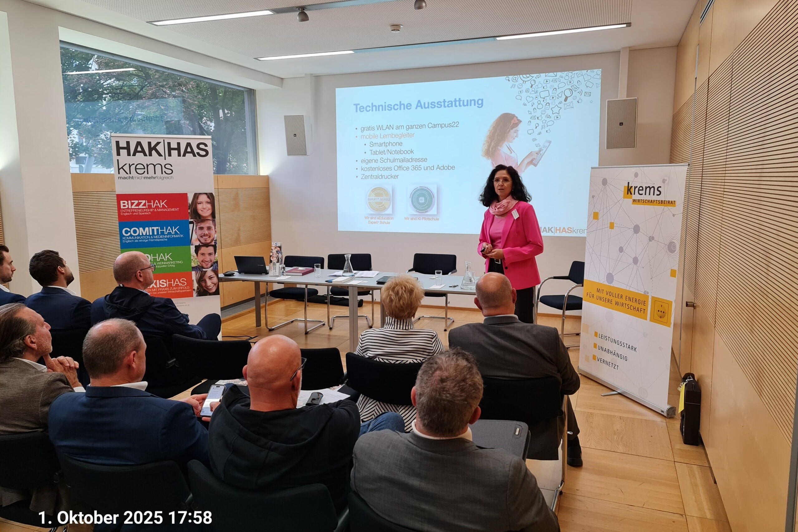 Vortrag vor 9 Personen in einem Seminarraum, eine Frau präsentiert vor einer Leinwand, Im Hintergrund sieht man ein Banner der HAK|Haskrems und deren zweige sowie ein 2ter Banner der Stadt Krems