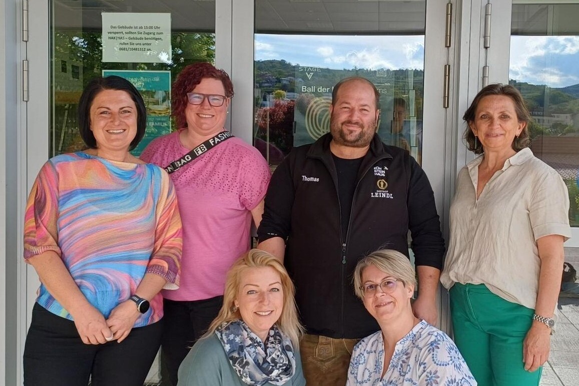 Gruppenfoto vor dem Eingang der HAK/HAS mit sechs Erwachsenen, die gemeinsam in die Kamera lächeln.