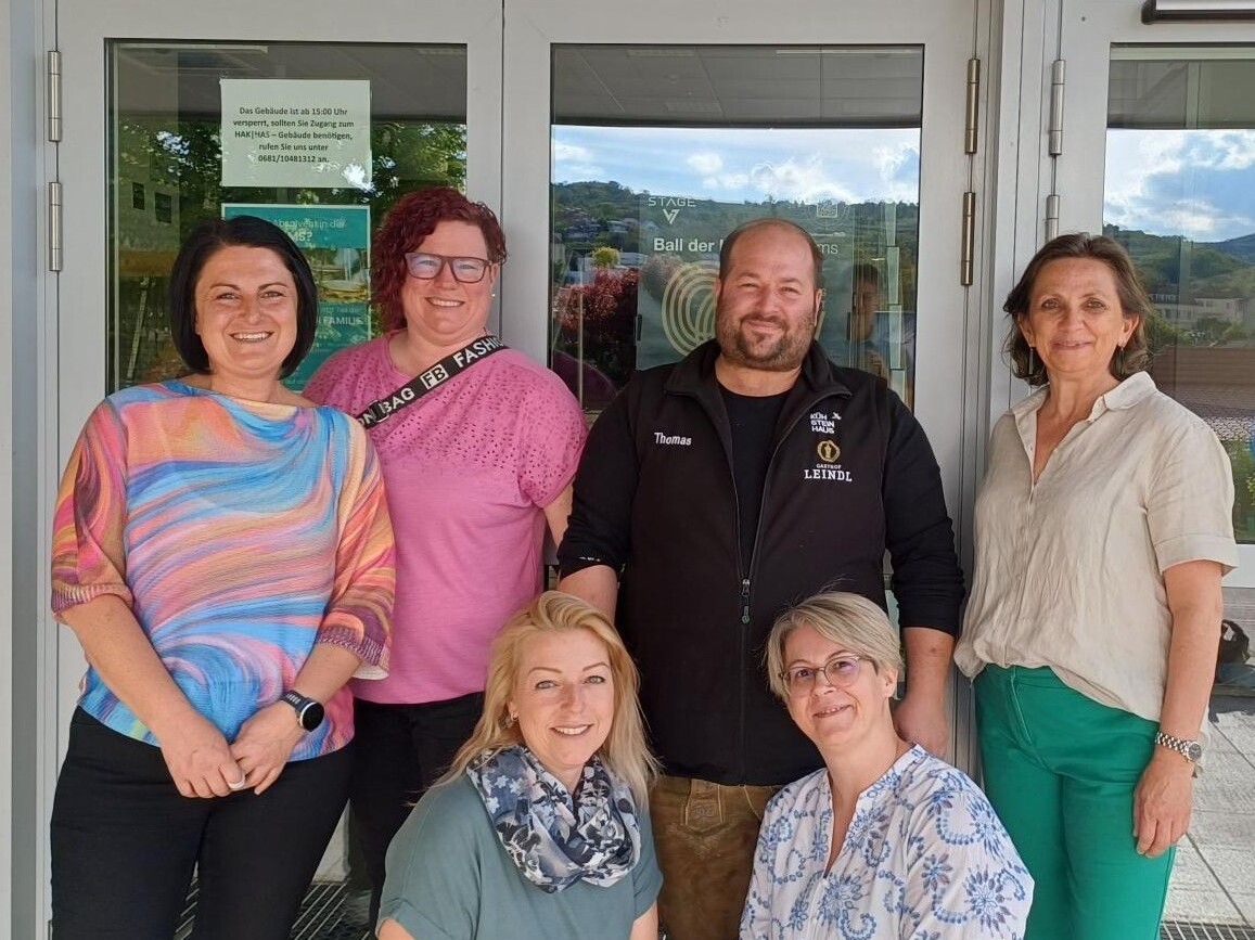 Gruppenfoto vor dem Eingang der HAK/HAS mit sechs Erwachsenen, die gemeinsam in die Kamera lächeln.