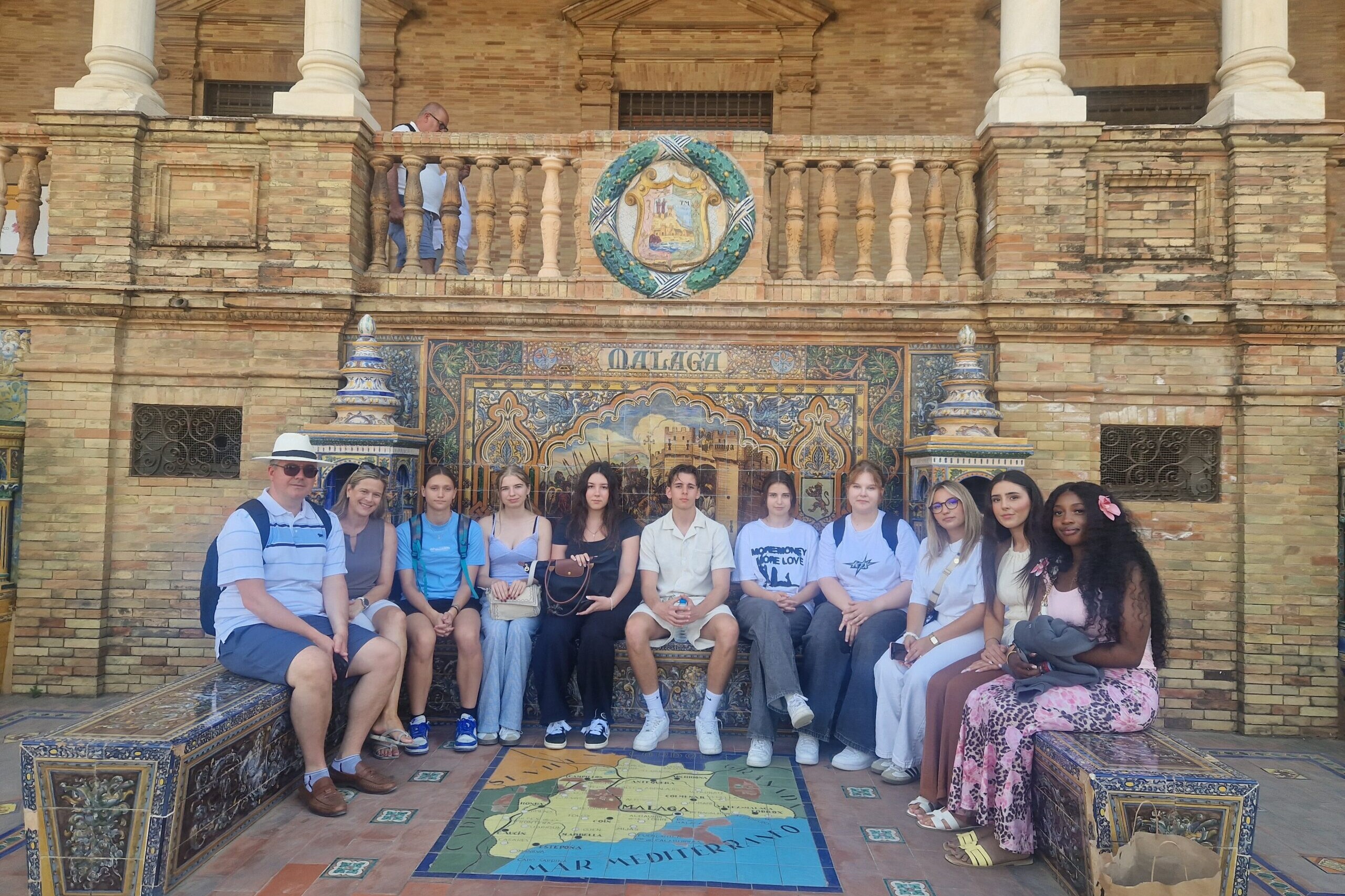 Ein Lehrer, eine Lehrerin und neun Schüler*innen auf einer Bank sitzend in die Kamera blickend.