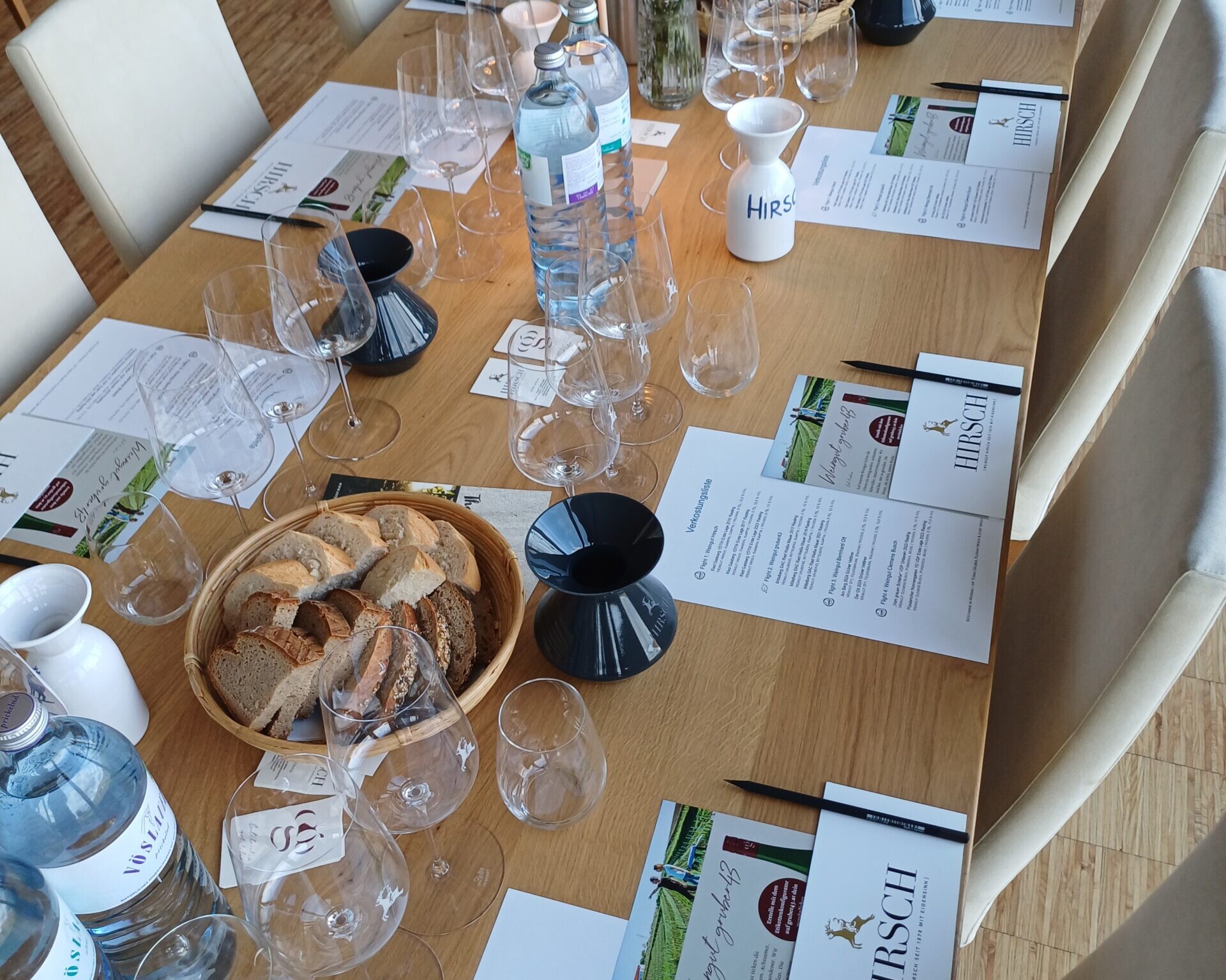 Gedeckter Tisch von oben ohne Gäste. Wasserflaschen, Weingläser, Brotkörbe und Verkostungsliste sind zu sehen.