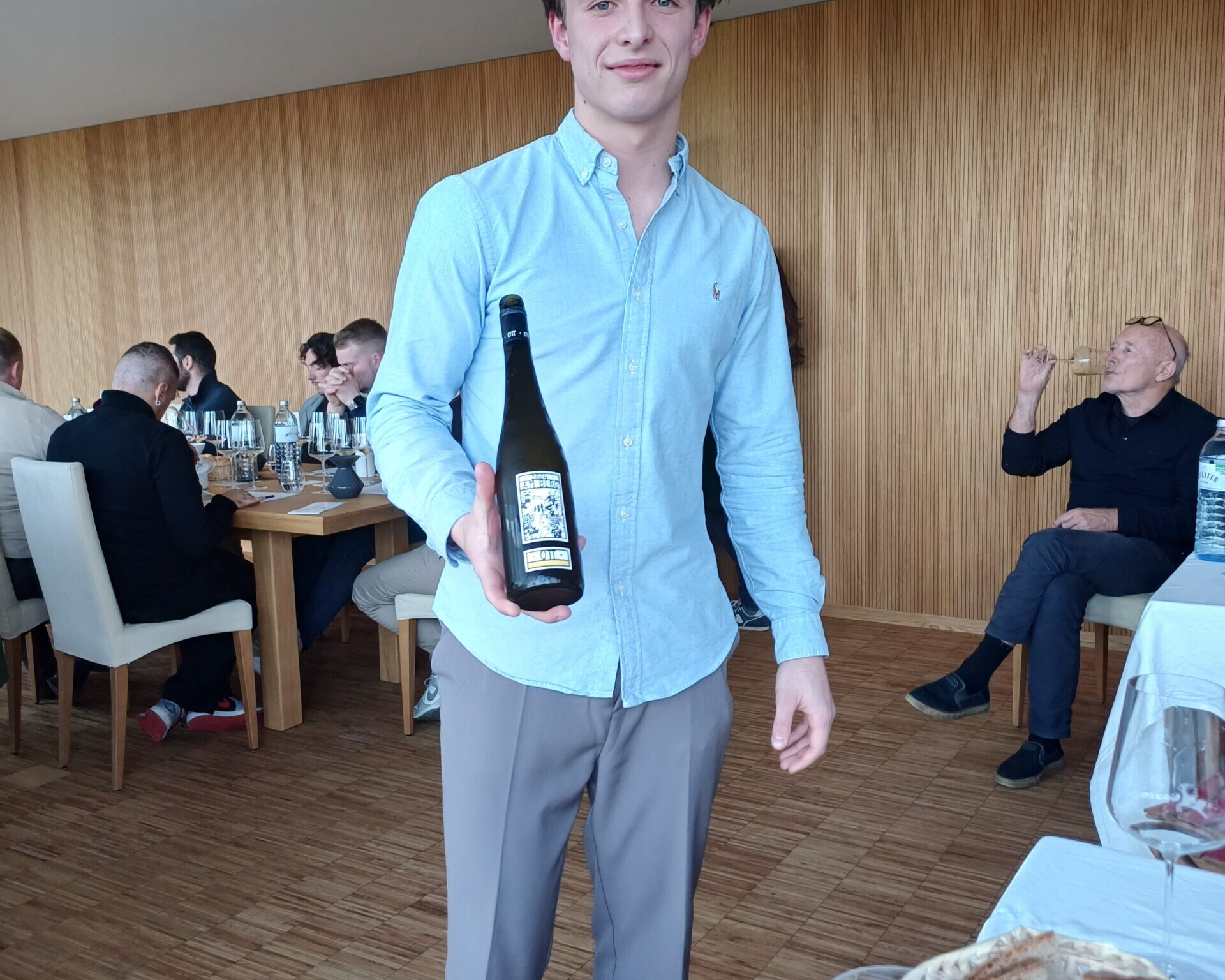 Schüler, direkt in die Kamera sehend, hält Weinflasche zeigend in der Hand. Weitere Gäste sind im Hintergrund.