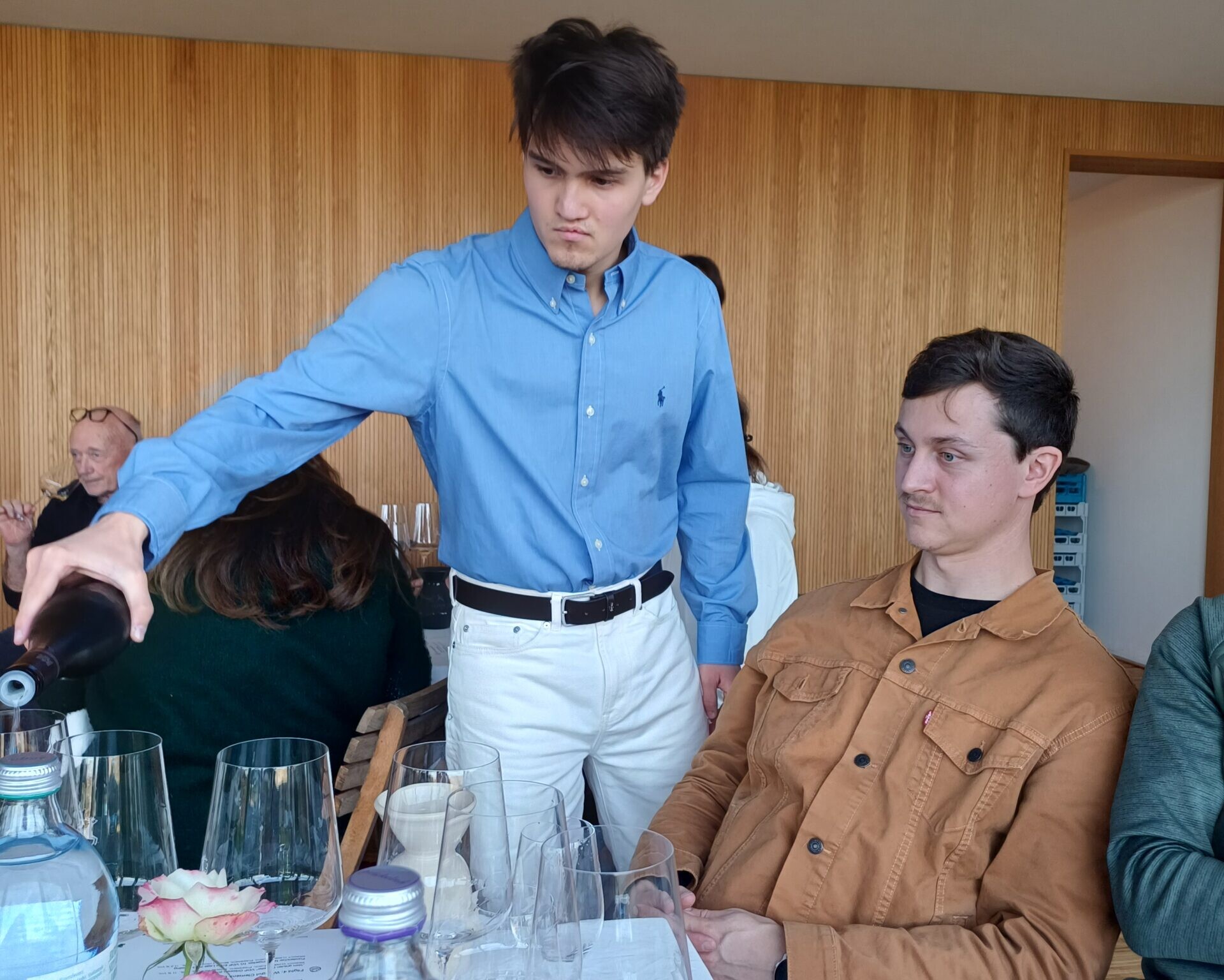 Schüler gießt Gast ein Glas Wein ein. Weitere Gäste im Hintergrund.