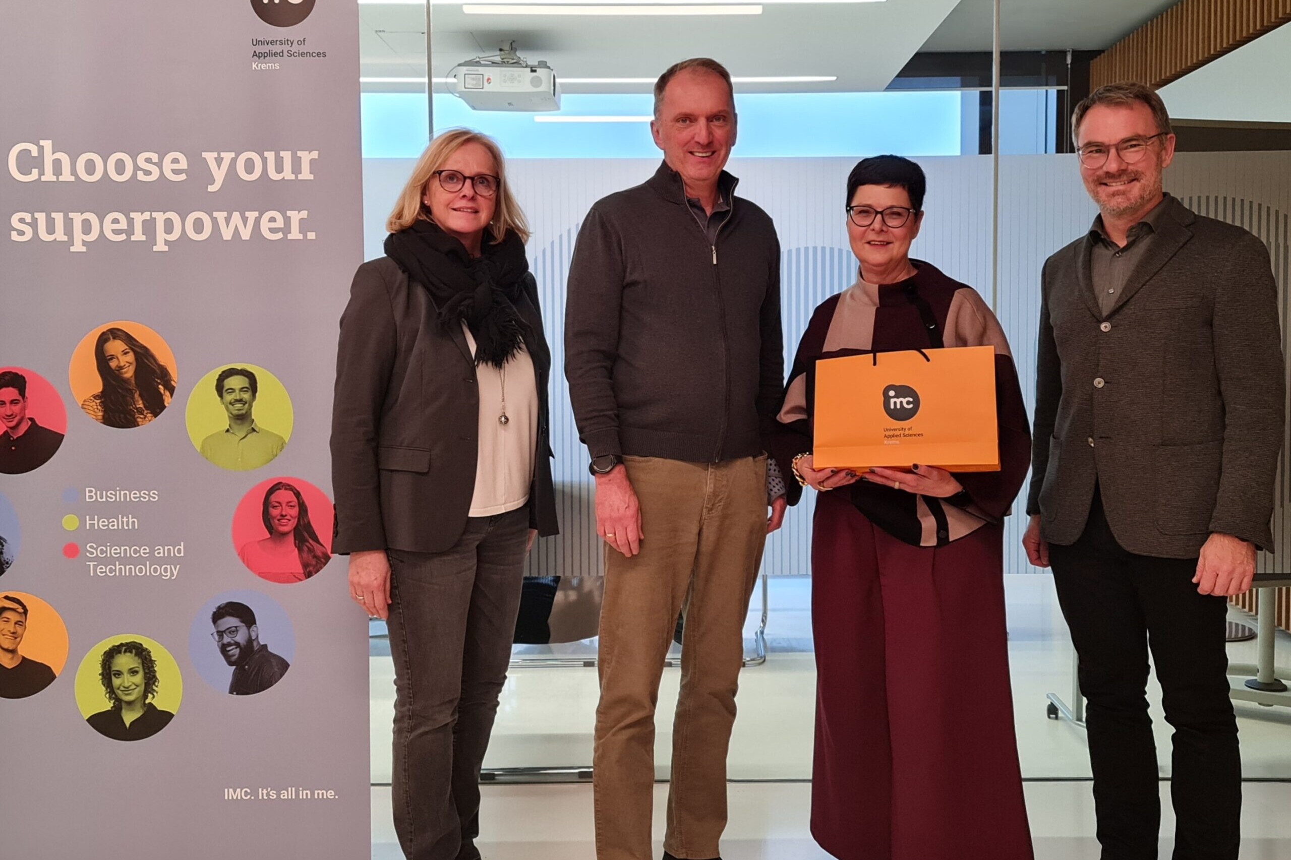 Direktorin, mit orangener IMC-Tasche in der Hand, gemeinsam mit Lehrer und zwei IMC-Professor*innen neben IMC-Roll-up.