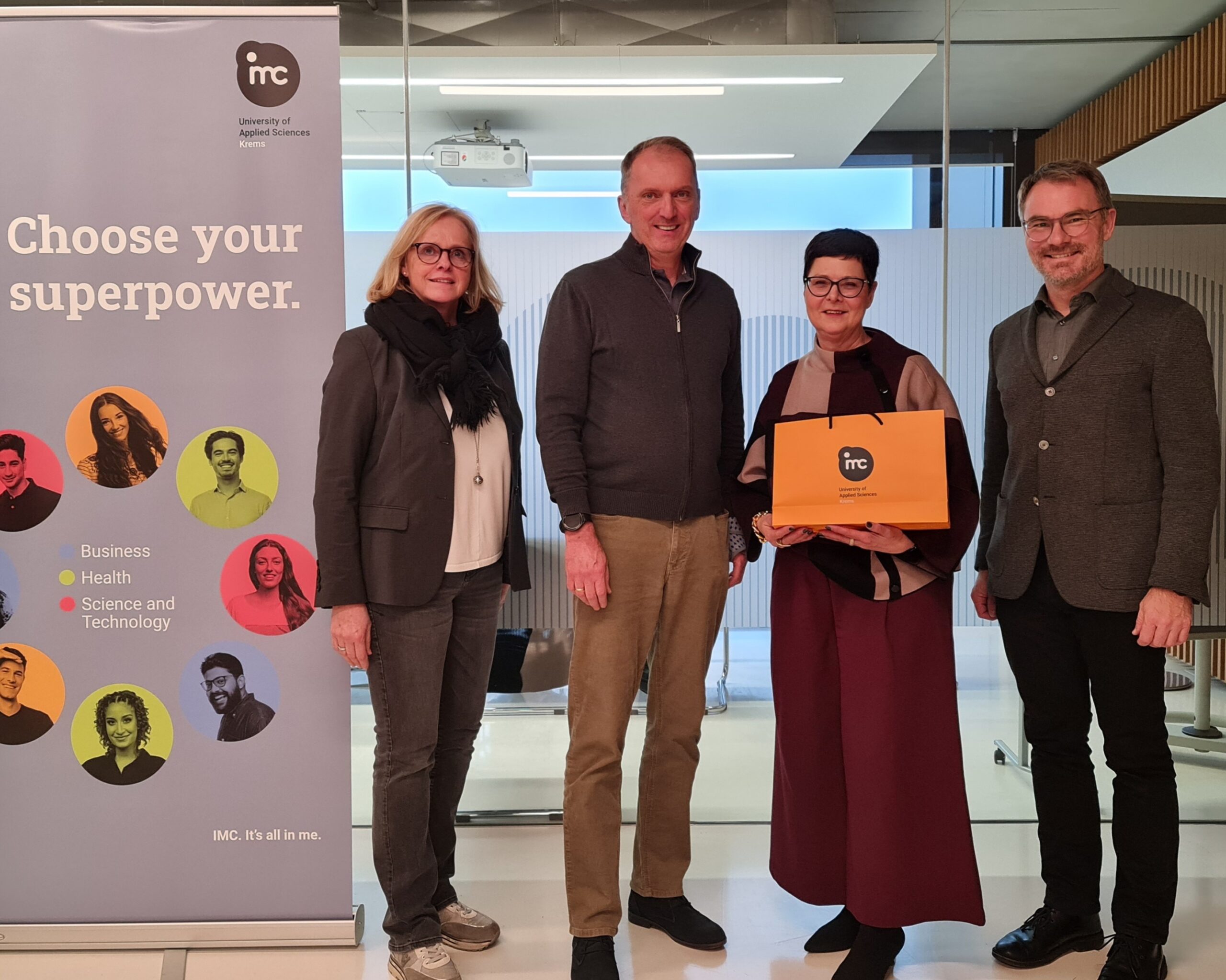 Direktorin, mit orangener IMC-Tasche in der Hand, gemeinsam mit Lehrer und zwei IMC-Professor*innen neben IMC-Roll-up.