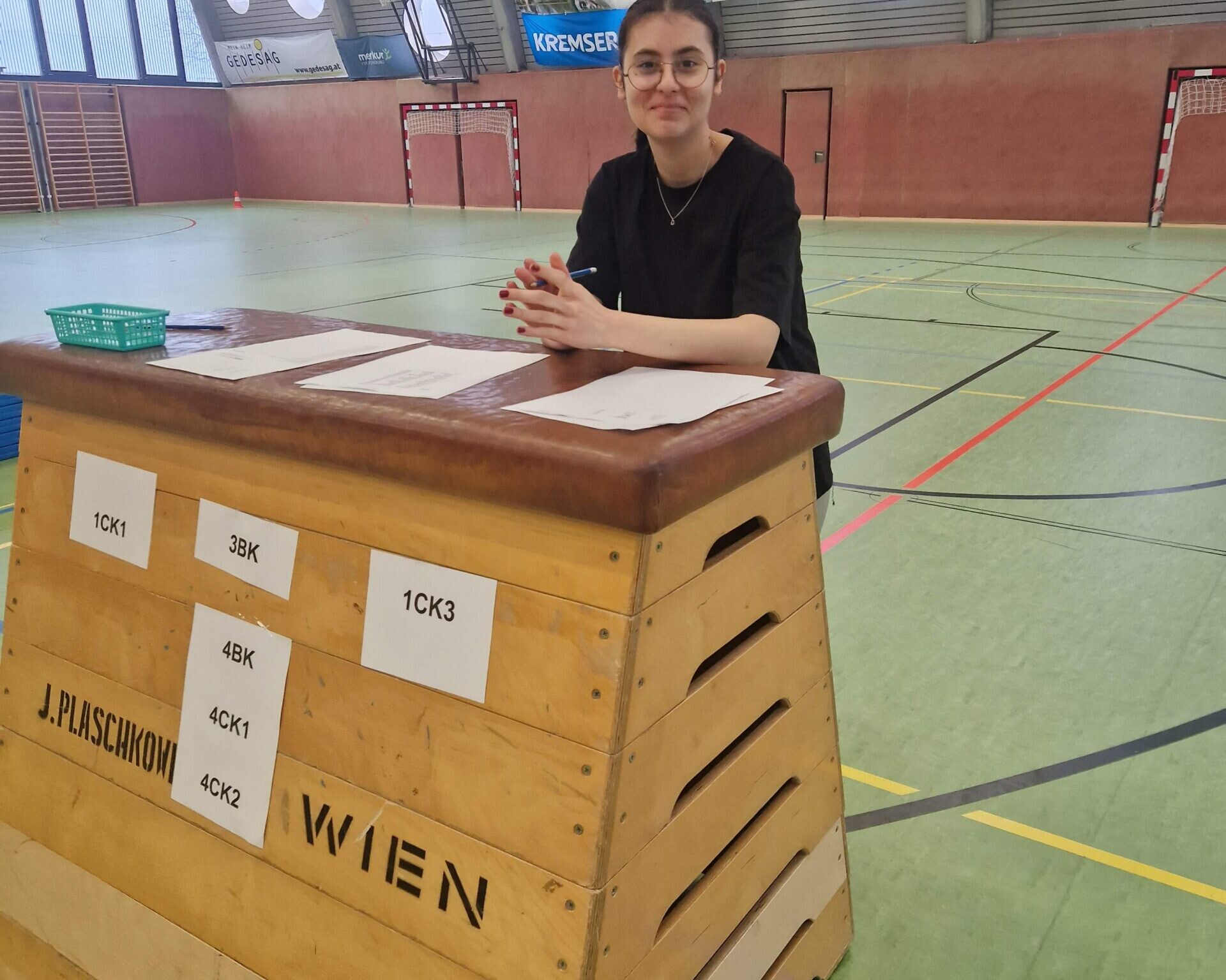 Schülerin stehend hinter Sprungkasten mit aufgeklebten Zetteln, direkt in die Kamera schauend.