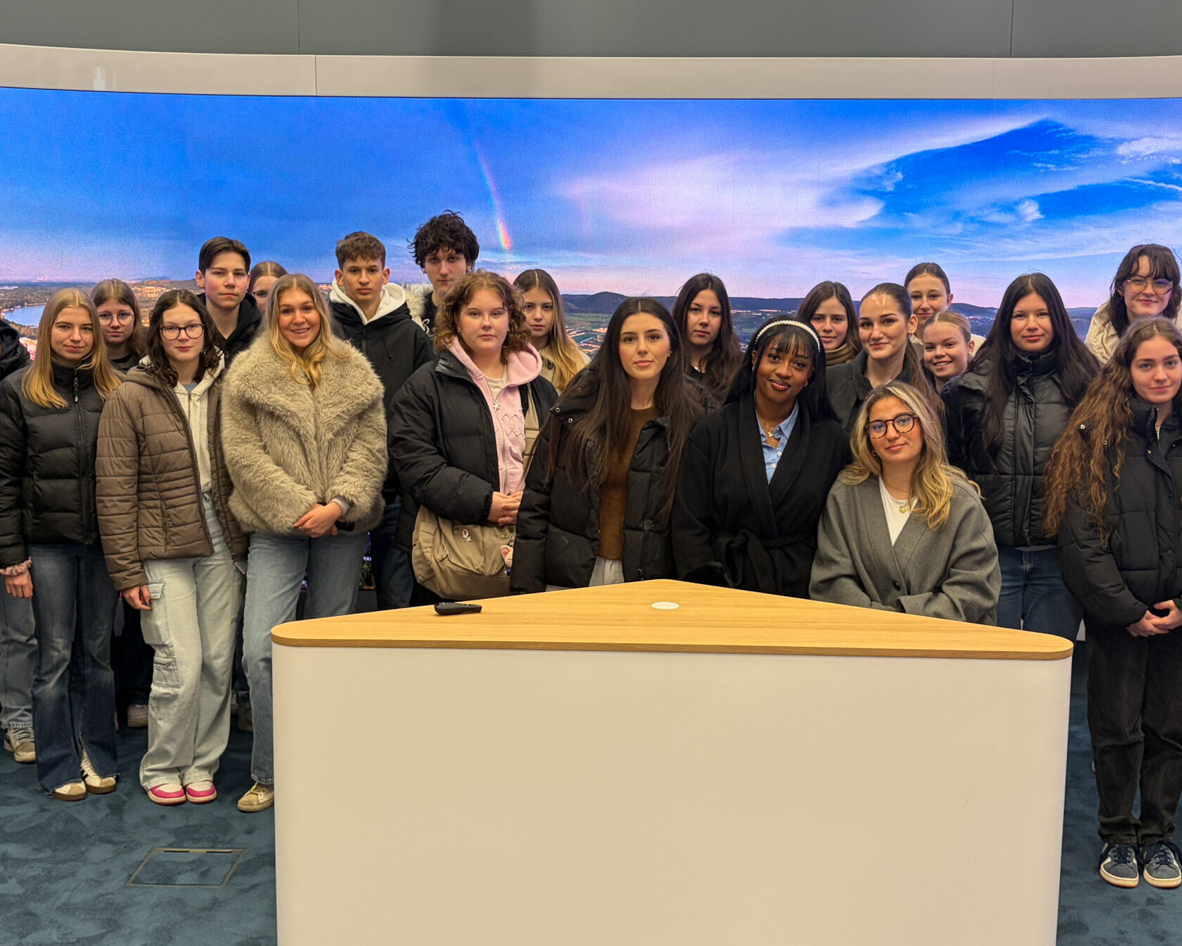 Eine Schülerklasse, eine Lehrerin und eine Betreuerin stehen hinter einem Nachrichten-Studio-Tisch.