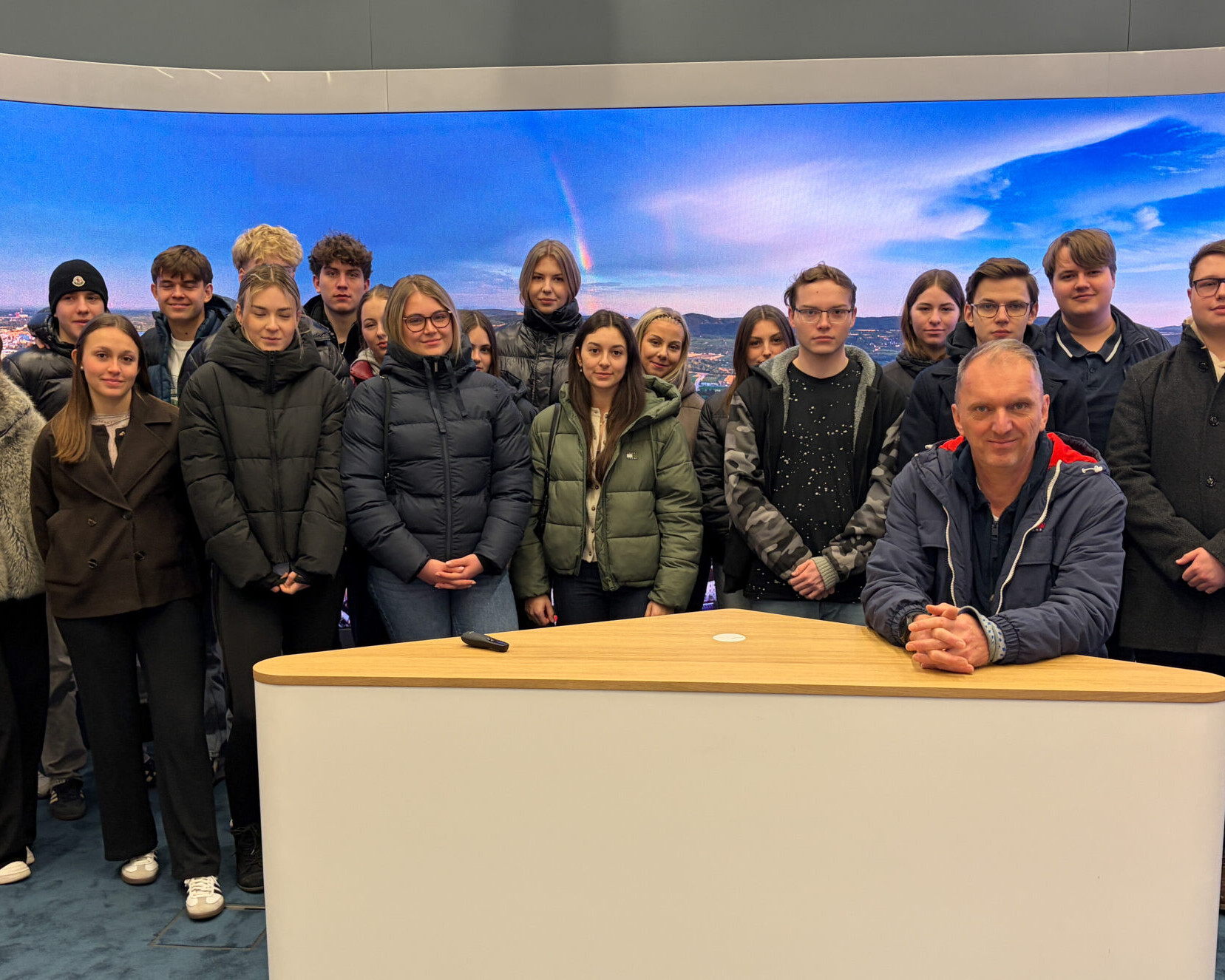 Eine Schülerklasse und ein Lehrer stehen hinter einem Nachrichten-Studio-Tisch.