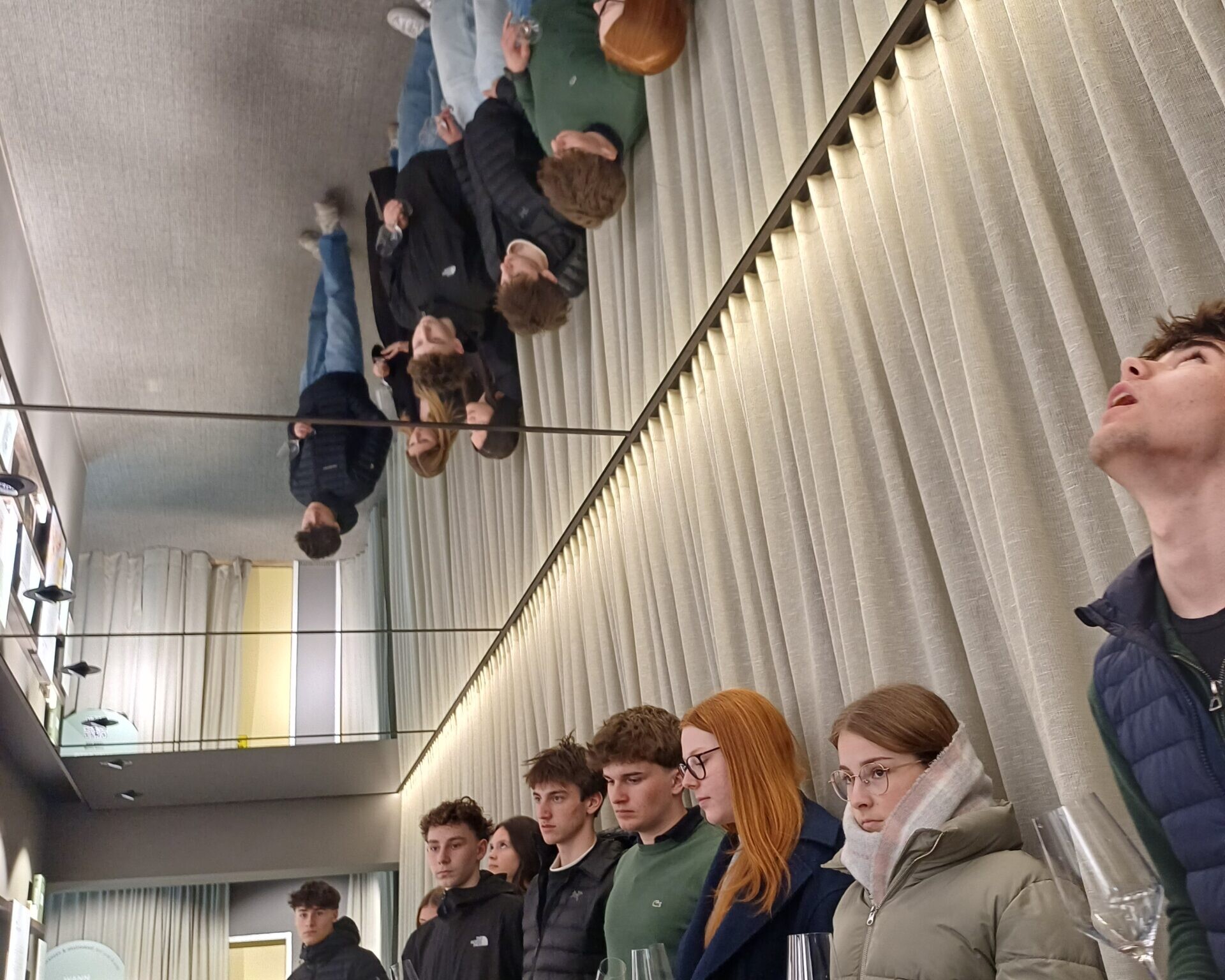 Schüler:innen stehen mit einem Weinglas in der Hand an einer Wand. Die Decke ober ihnen ist ein Spiegel.