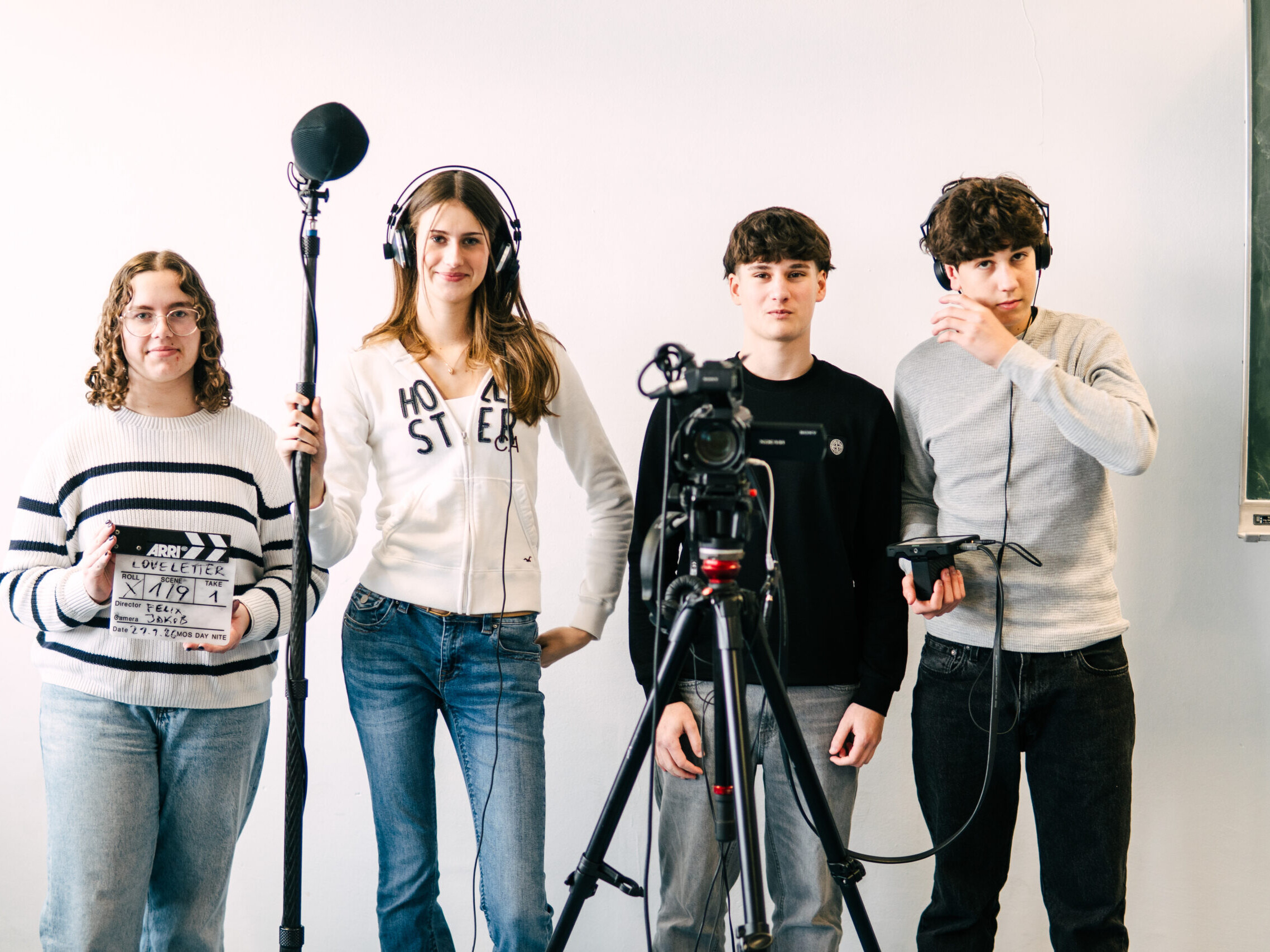 Vier junge Schüler*innen stehen vor einer Kameraaufstellung: eine Person hält eine Filmklappe, eine hält eine Tonangel, und zwei bedienen Kamera- und Tonequipment.