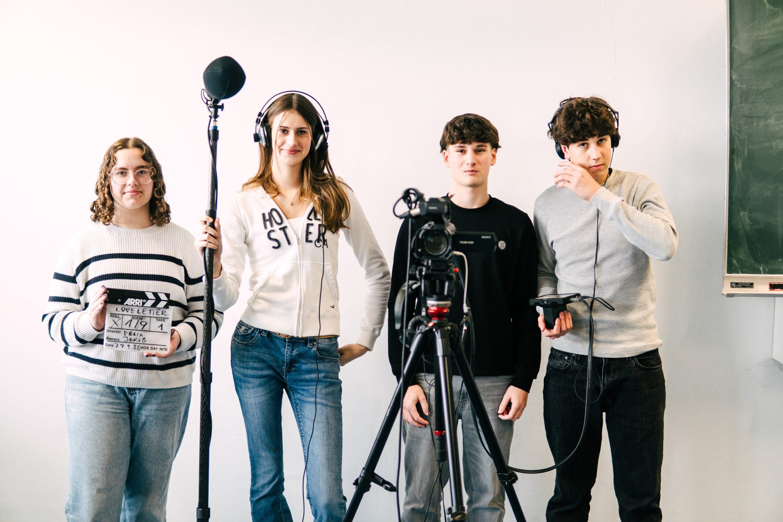 Vier junge Schüler*innen stehen vor einer Kameraaufstellung: eine Person hält eine Filmklappe, eine hält eine Tonangel, und zwei bedienen Kamera- und Tonequipment.