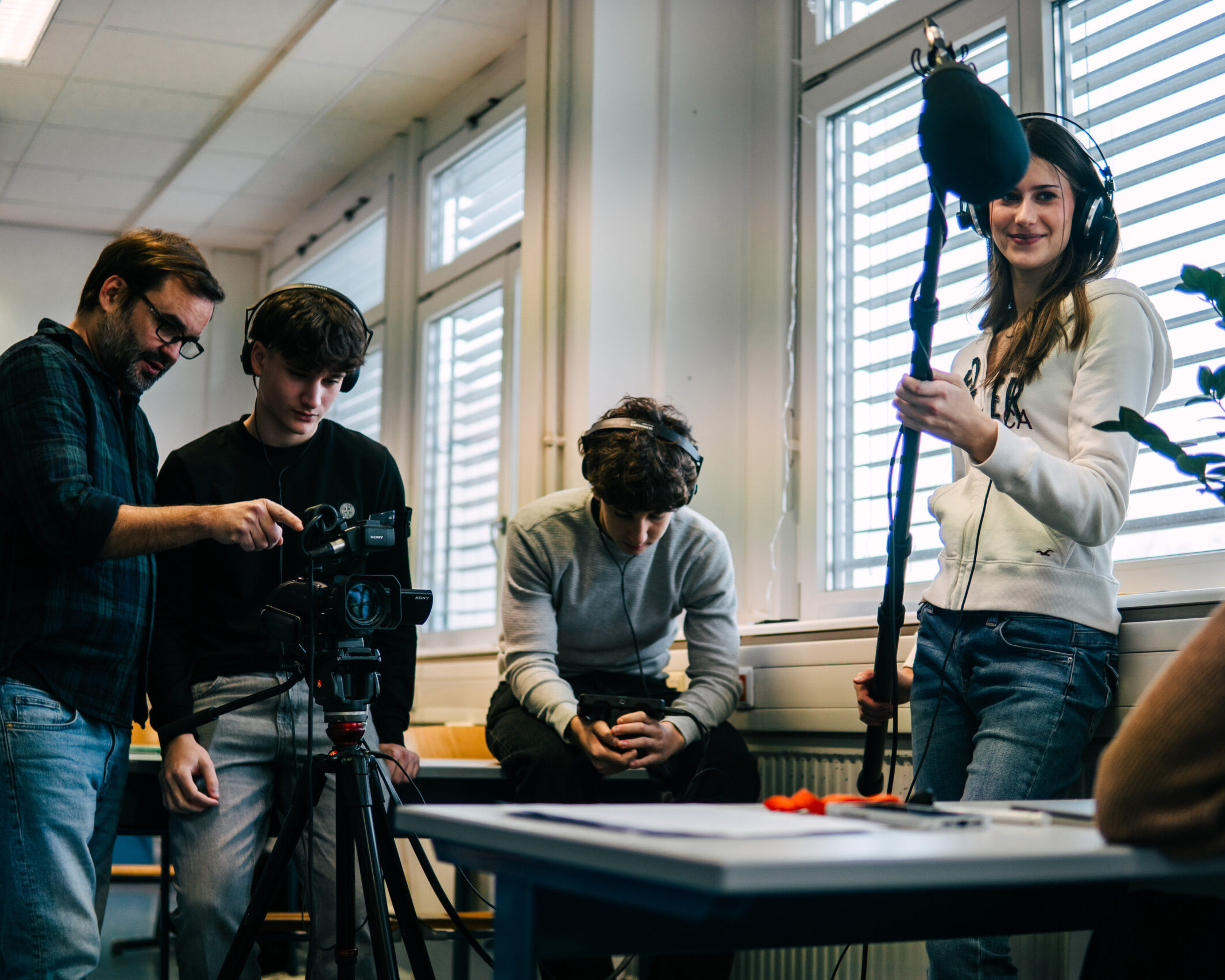 Mehrere junge Schüler*innen arbeiten in einem Klassenraum gemeinsam an einer Filmaufnahme – eine Person bedient die Kamera, eine andere hält eine Tonangel, während weitere daneben stehen oder sitzen.