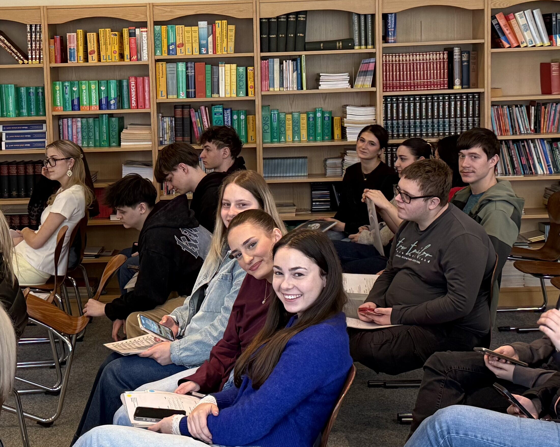 Gruppe von Schüler:innen sitzt in einem Klassenraum vor Bücherregalen und lächelt in die Kamera.