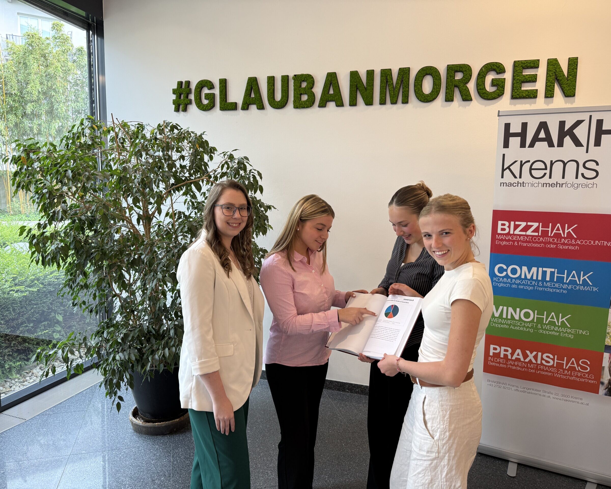 3 Schülerinnen und eine Frau stehen vor einer Wand mit der Aufschrift #GLAUBANMORGEN, rechts von ihnen ein Banner der HAK|Haskrems