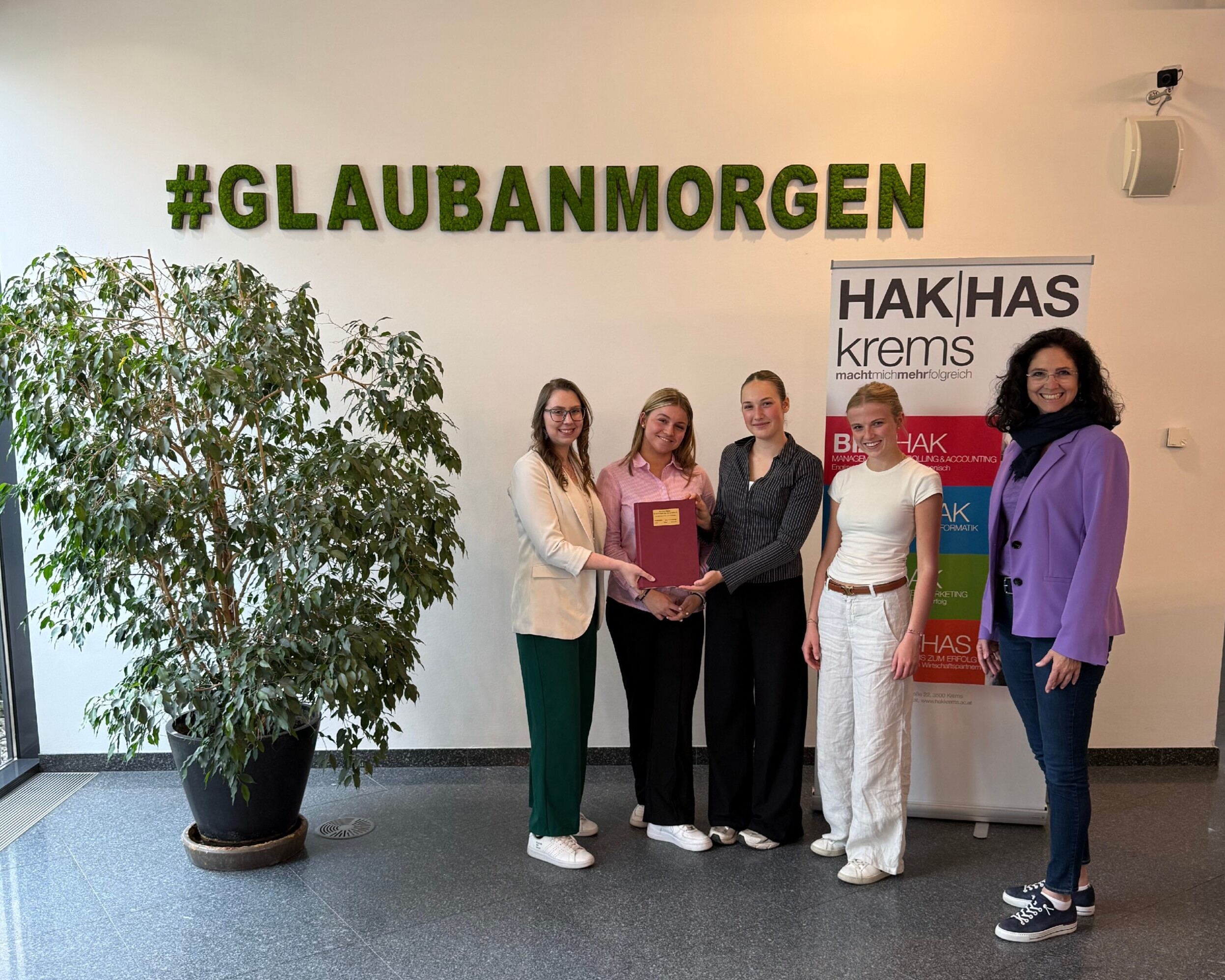 3 Schülerinnen, eine Lehrerin und eine Frau stehen vor einer Wand mit der Aufschrift #GLAUBANMORGEN, rechts von ihnen ein Banner der HAK|Haskrems