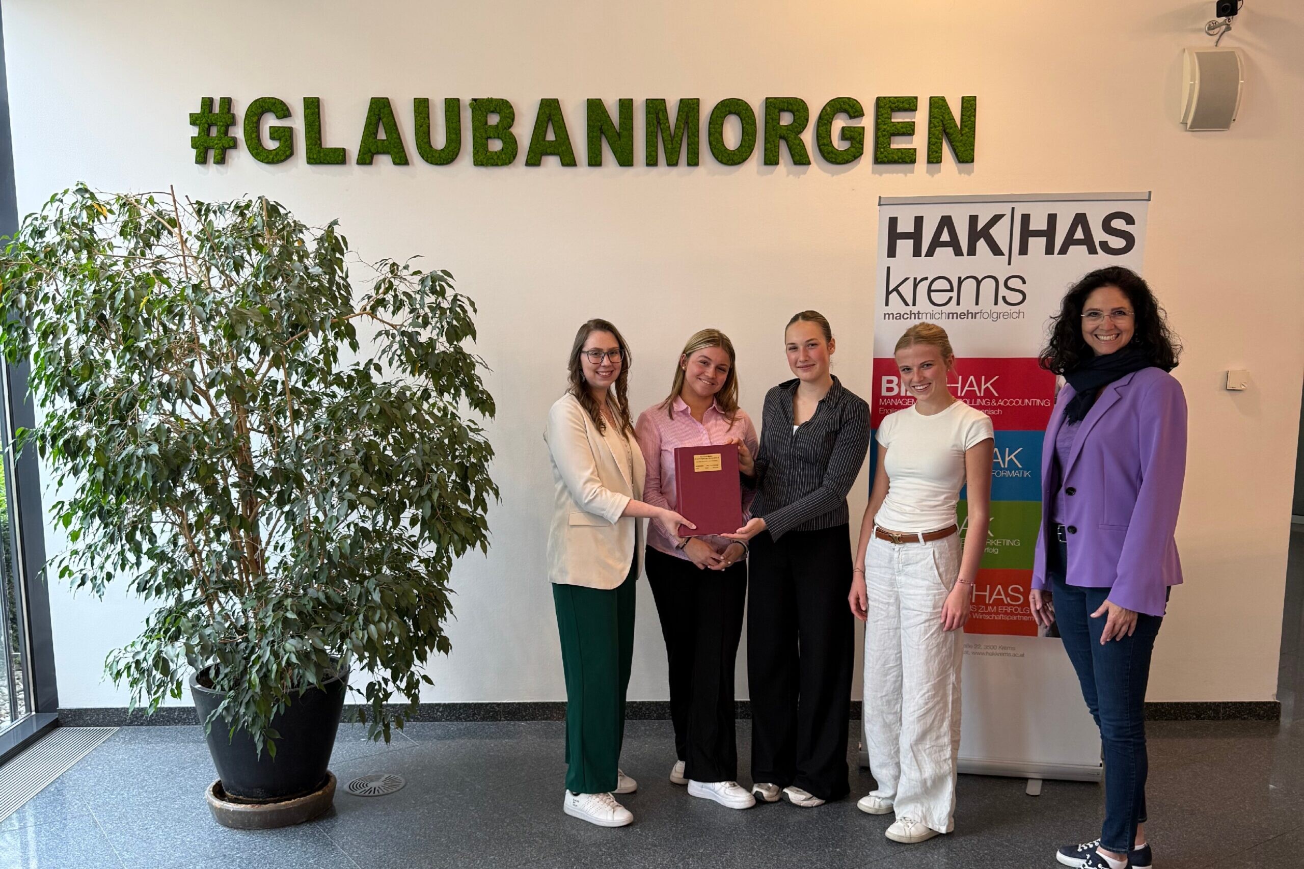 3 Schülerinnen, eine Lehrerin und eine Frau stehen vor einer Wand mit der Aufschrift #GLAUBANMORGEN, rechts von ihnen ein Banner der HAK|Haskrems