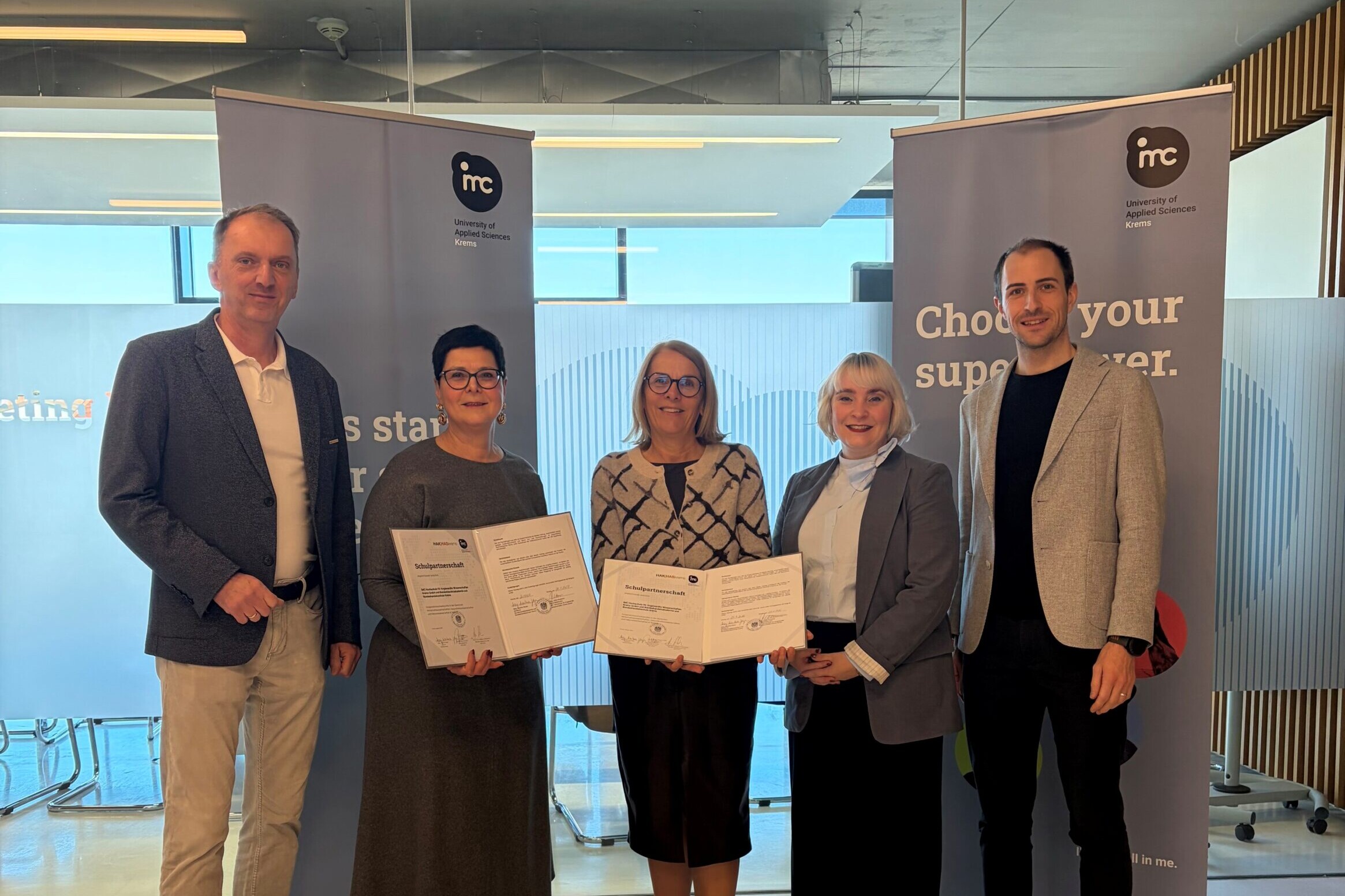 Fünf Erwachsene stehen bei einer Zertifikatsübergabe vor Roll-up-Bannern der IMC Fachhochschule Krems und halten Urkunden in der Hand.