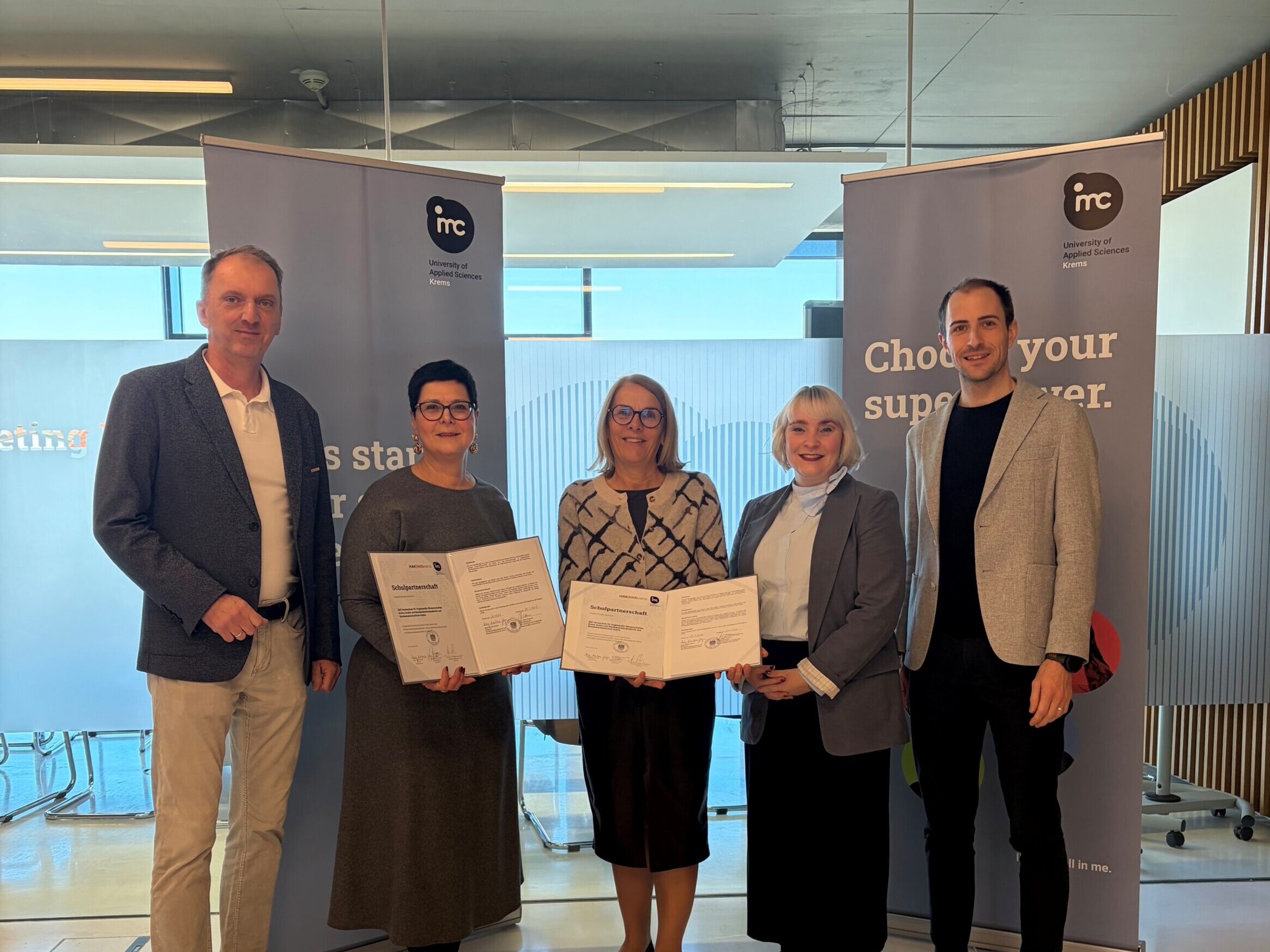 Fünf Erwachsene stehen bei einer Zertifikatsübergabe vor Roll-up-Bannern der IMC Fachhochschule Krems und halten Urkunden in der Hand.