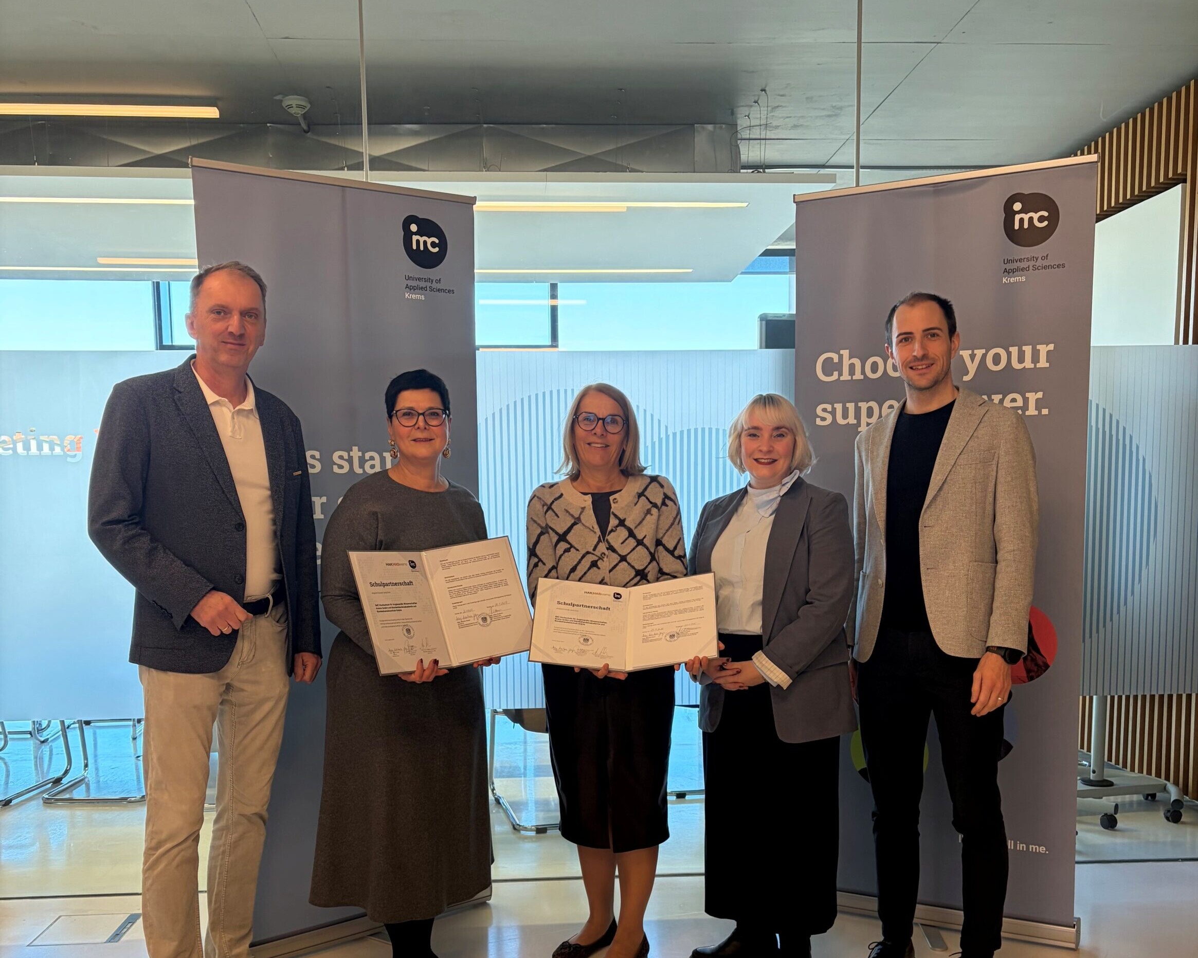 Fünf Erwachsene stehen bei einer Zertifikatsübergabe vor Roll-up-Bannern der IMC Fachhochschule Krems und halten Urkunden in der Hand.