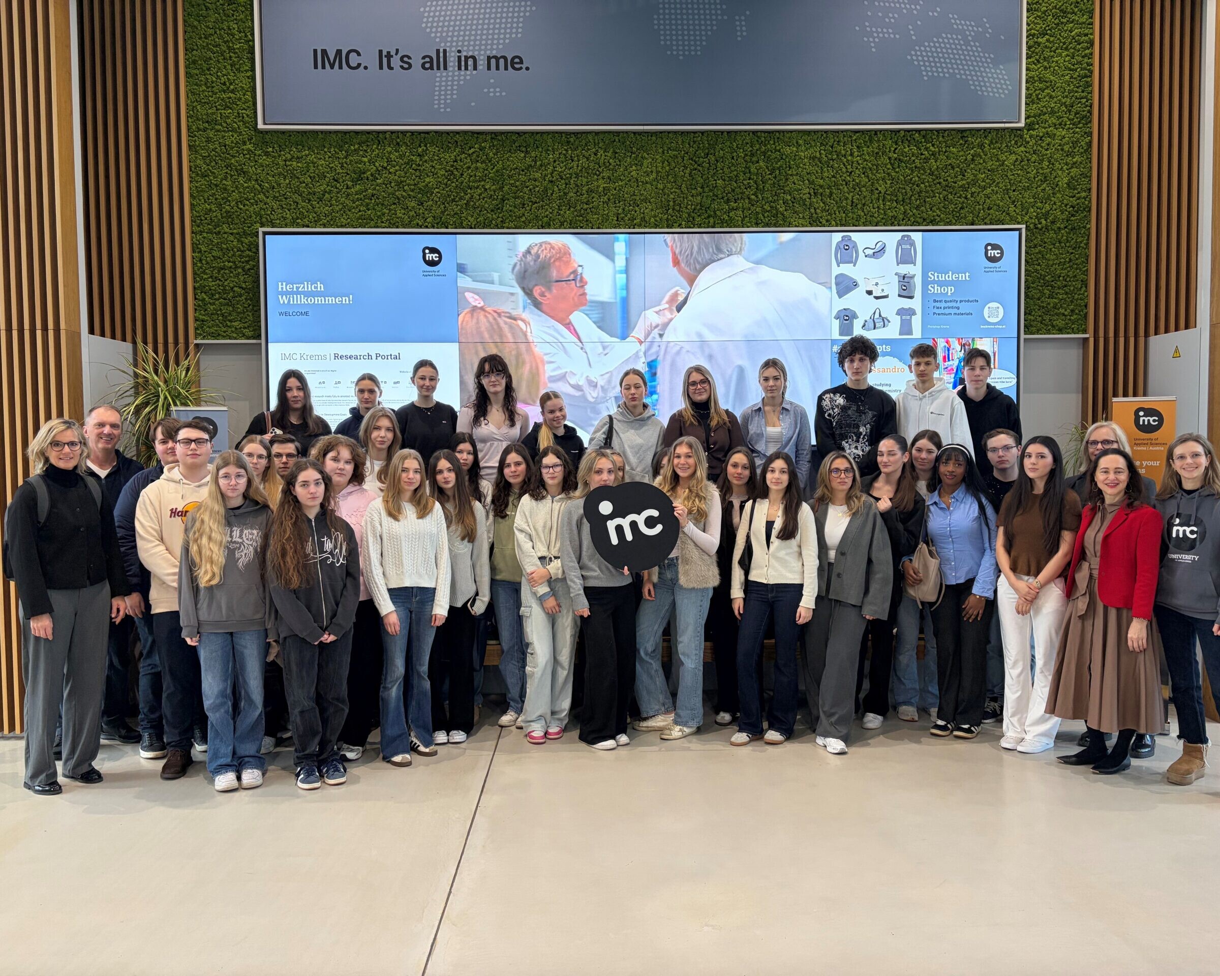 Zwei Lehrer*innen, eine Betreuerin und viele Schüler*innen machen ein Gruppenfoto, in der Mitte hält eine Schülerin das IMC Krems Logo.