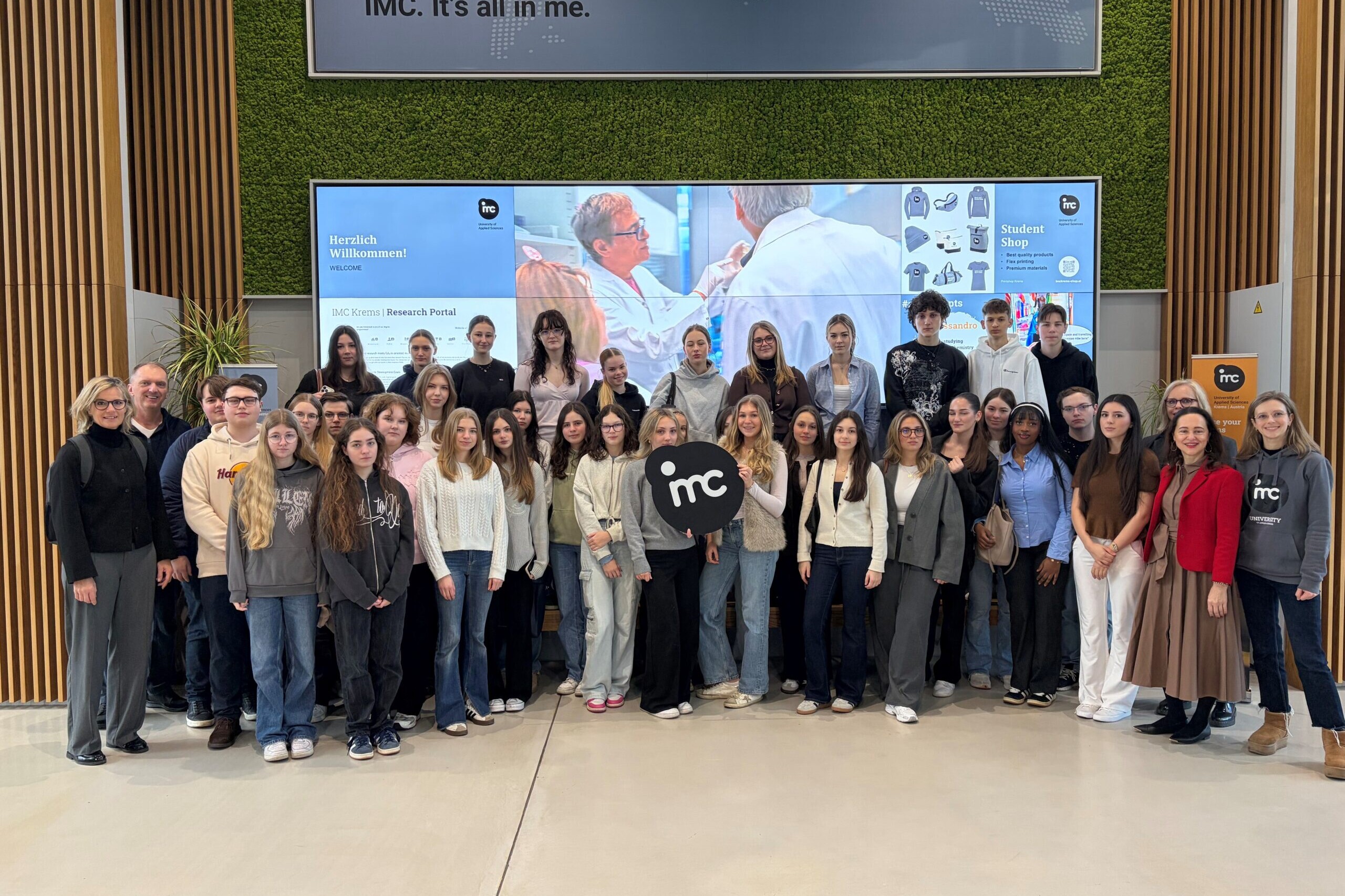 Zwei Lehrer*innen, eine Betreuerin und viele Schüler*innen machen ein Gruppenfoto, in der Mitte hält eine Schülerin das IMC Krems Logo.