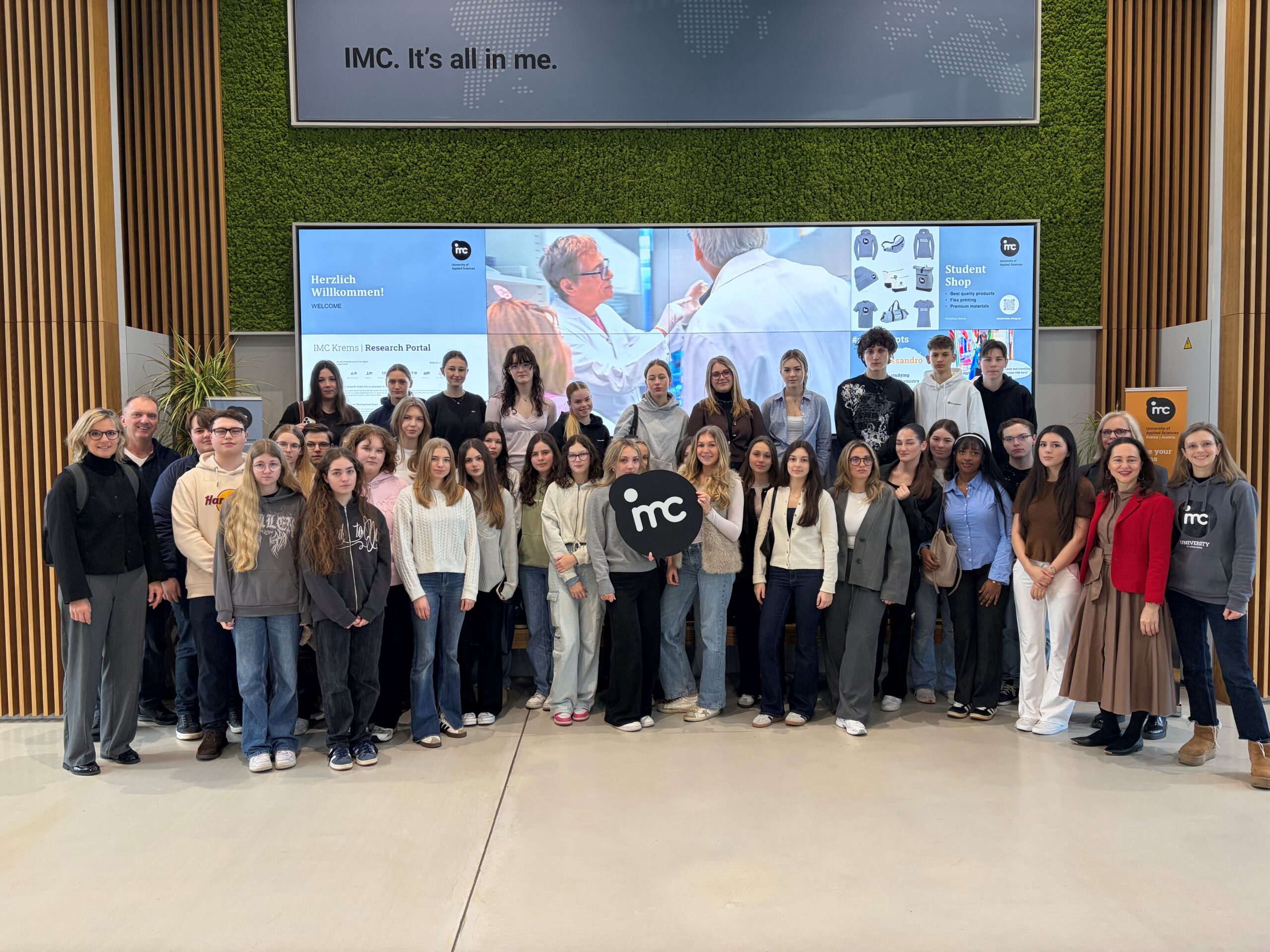 Zwei Lehrer*innen, eine Betreuerin und viele Schüler*innen machen ein Gruppenfoto, in der Mitte hält eine Schülerin das IMC Krems Logo.