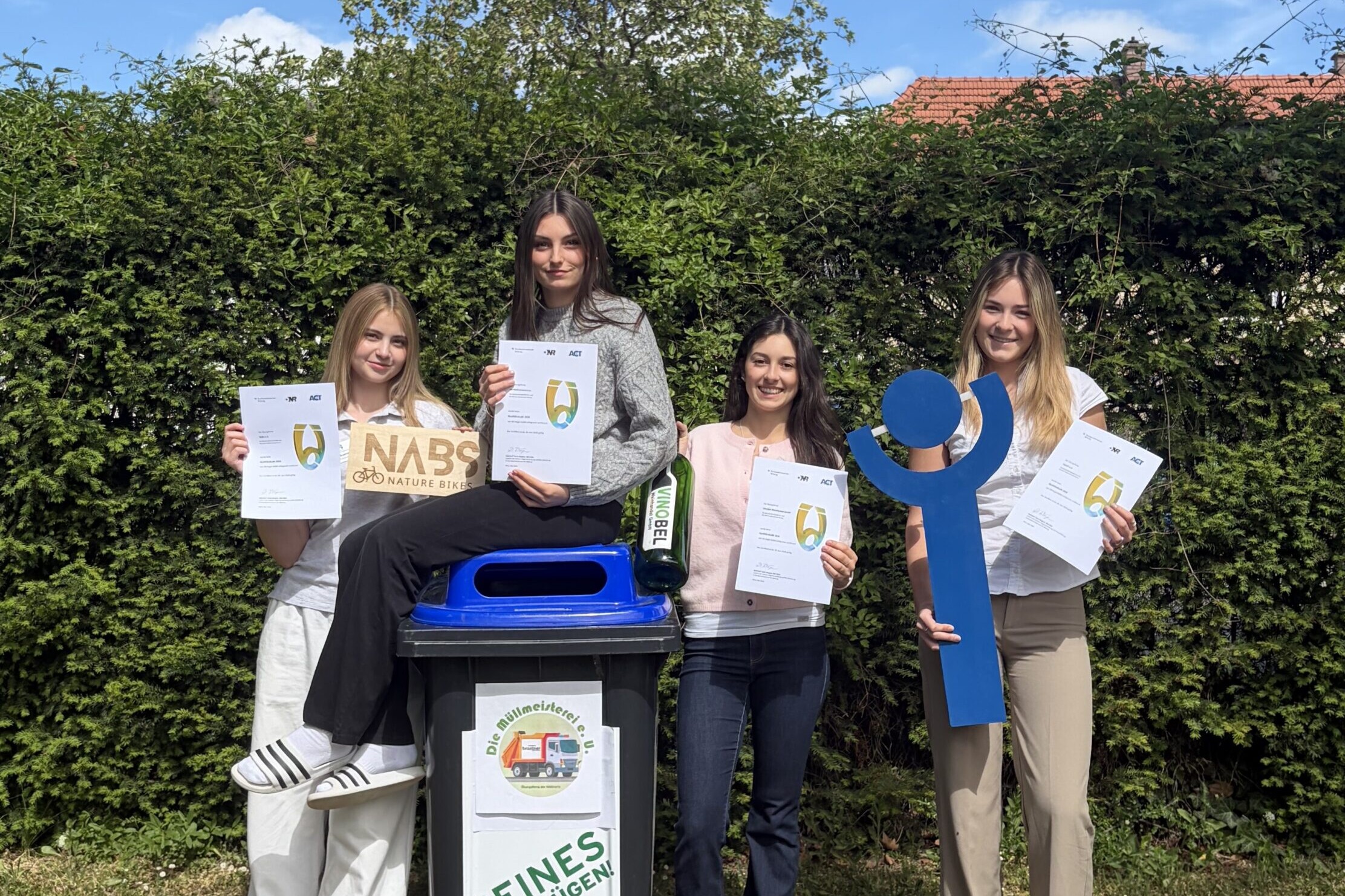 Vier Schülerinnen stehen draußen neben einer Mülltonne und halten Urkunden sowie Recycling Symbole in die Kamera.