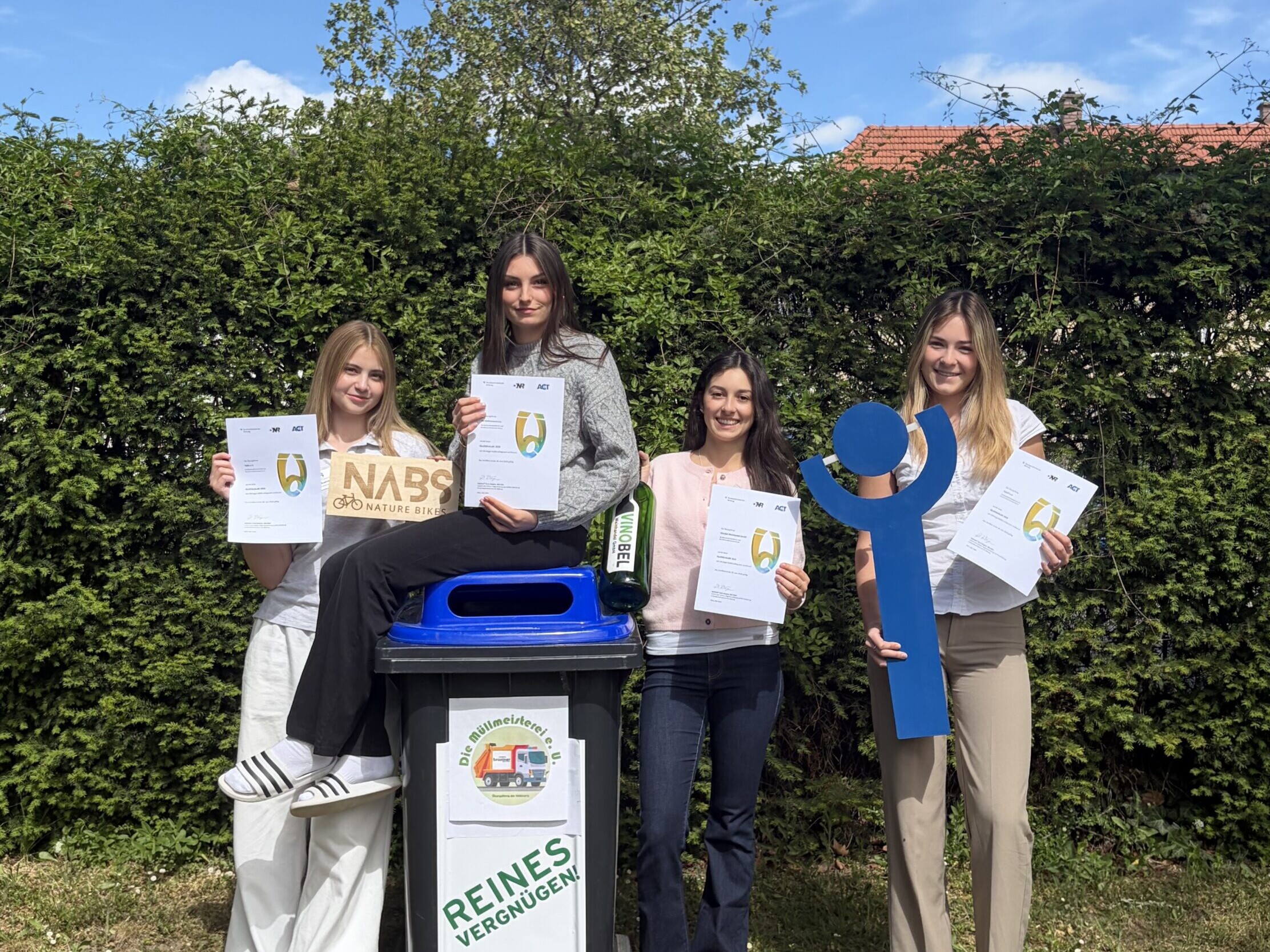 Vier Schülerinnen stehen draußen neben einer Mülltonne und halten Urkunden sowie Recycling Symbole in die Kamera.