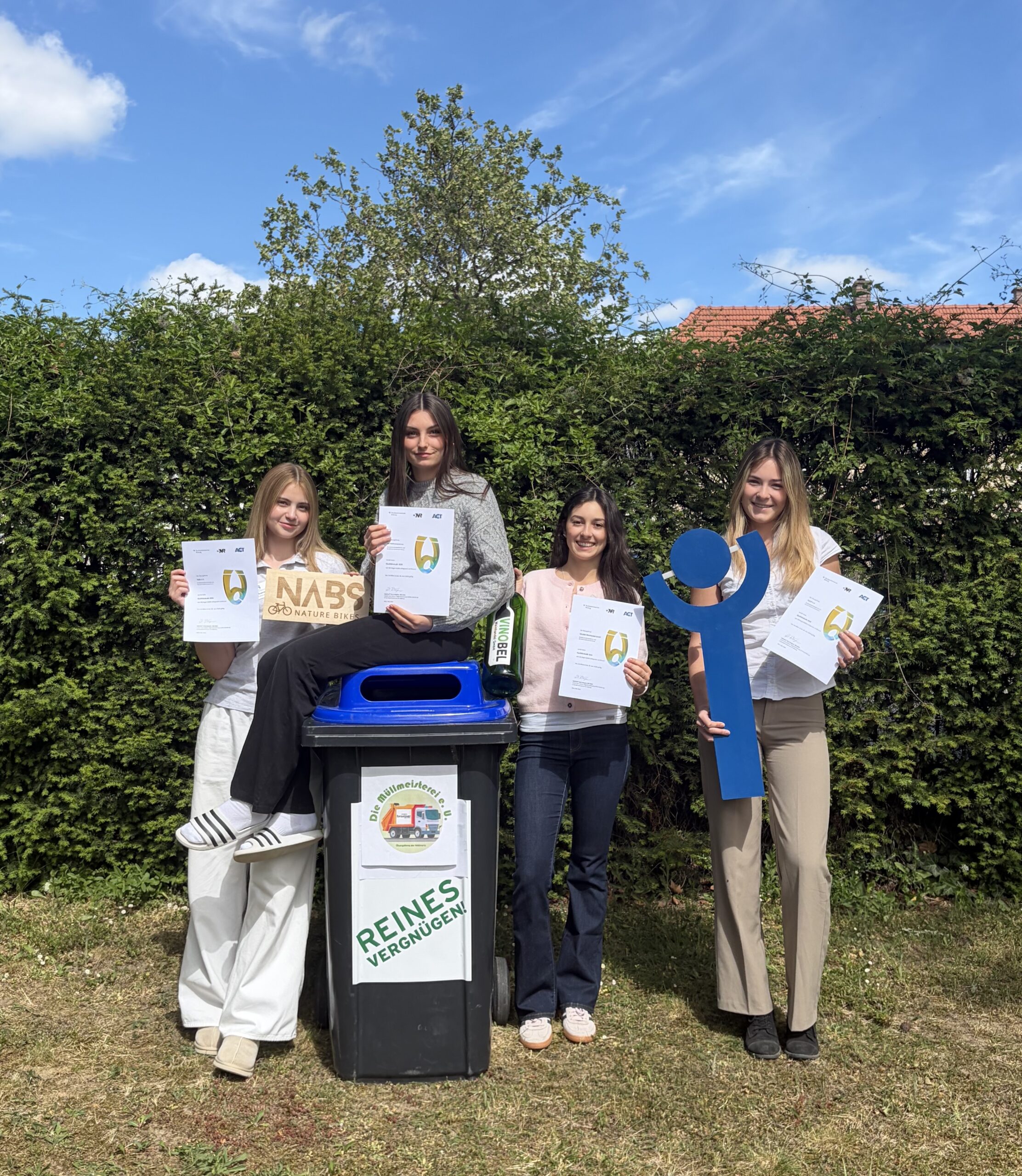 Vier Schülerinnen stehen draußen neben einer Mülltonne und halten Urkunden sowie Recycling Symbole in die Kamera.