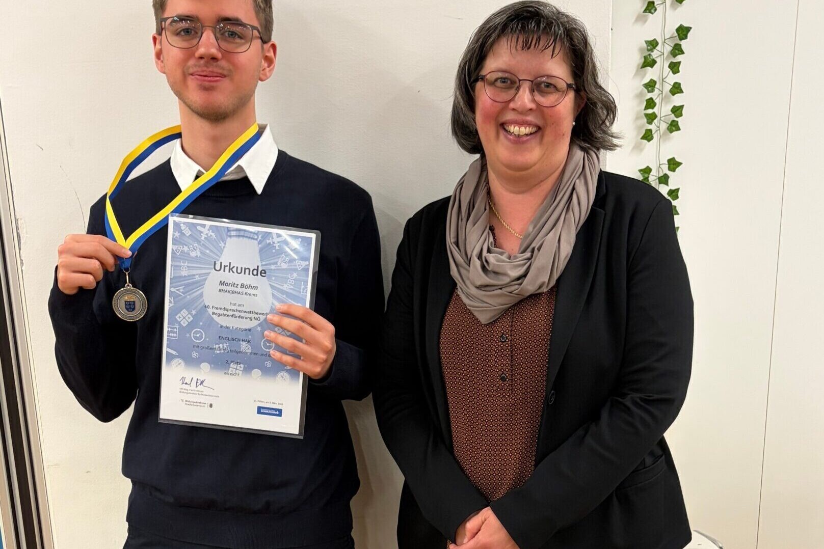 Eine Lehrerin und ein Schüler beim Fremdsprachenwettbewerb. Der Schüler hält eine Medaille und eine Urkunde.