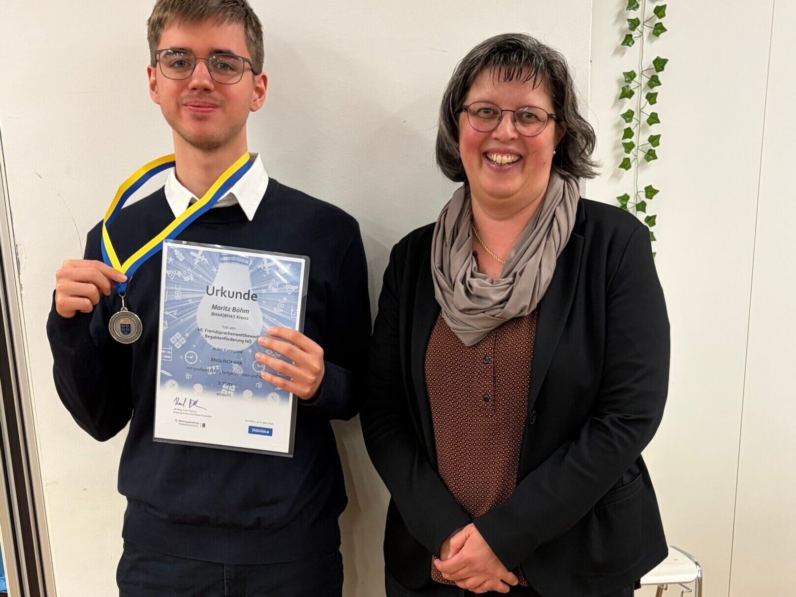 Eine Lehrerin und ein Schüler beim Fremdsprachenwettbewerb. Der Schüler hält eine Medaille und eine Urkunde.