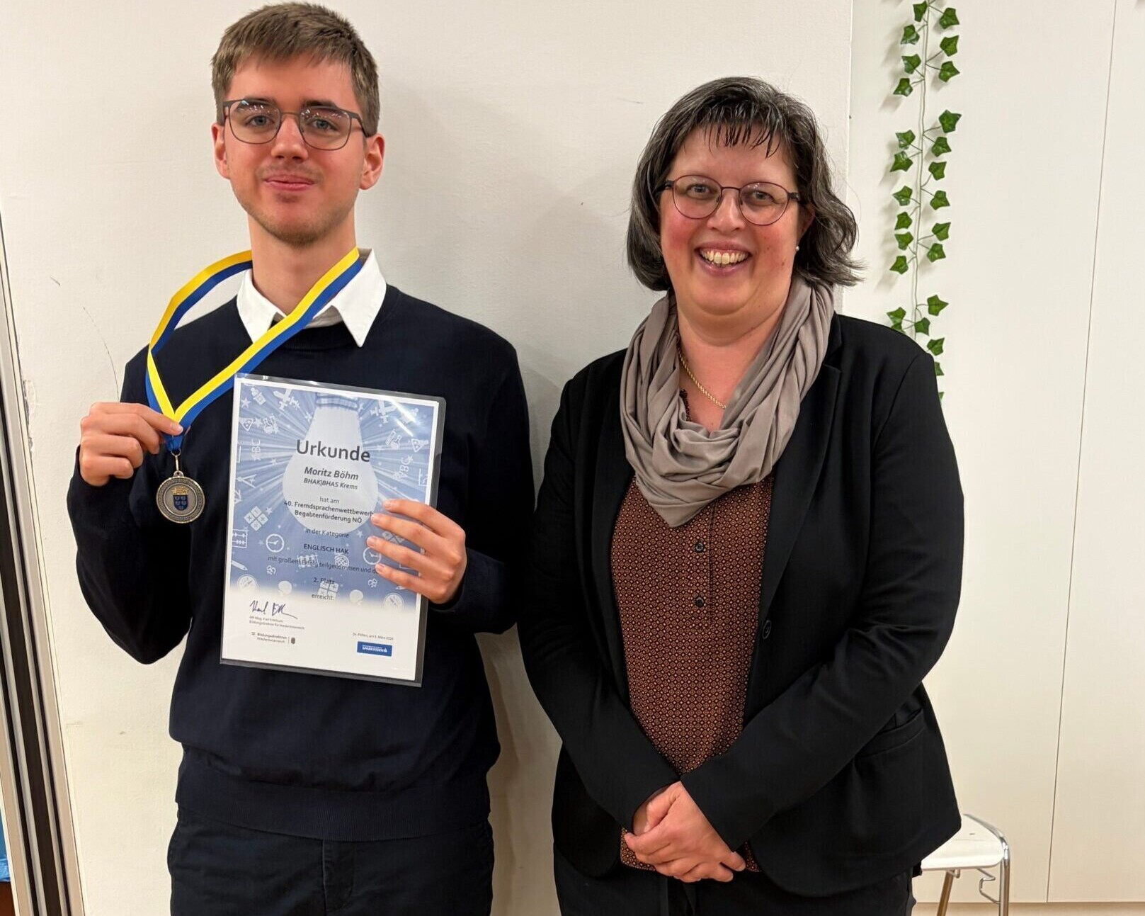 Eine Lehrerin und ein Schüler beim Fremdsprachenwettbewerb. Der Schüler hält eine Medaille und eine Urkunde.