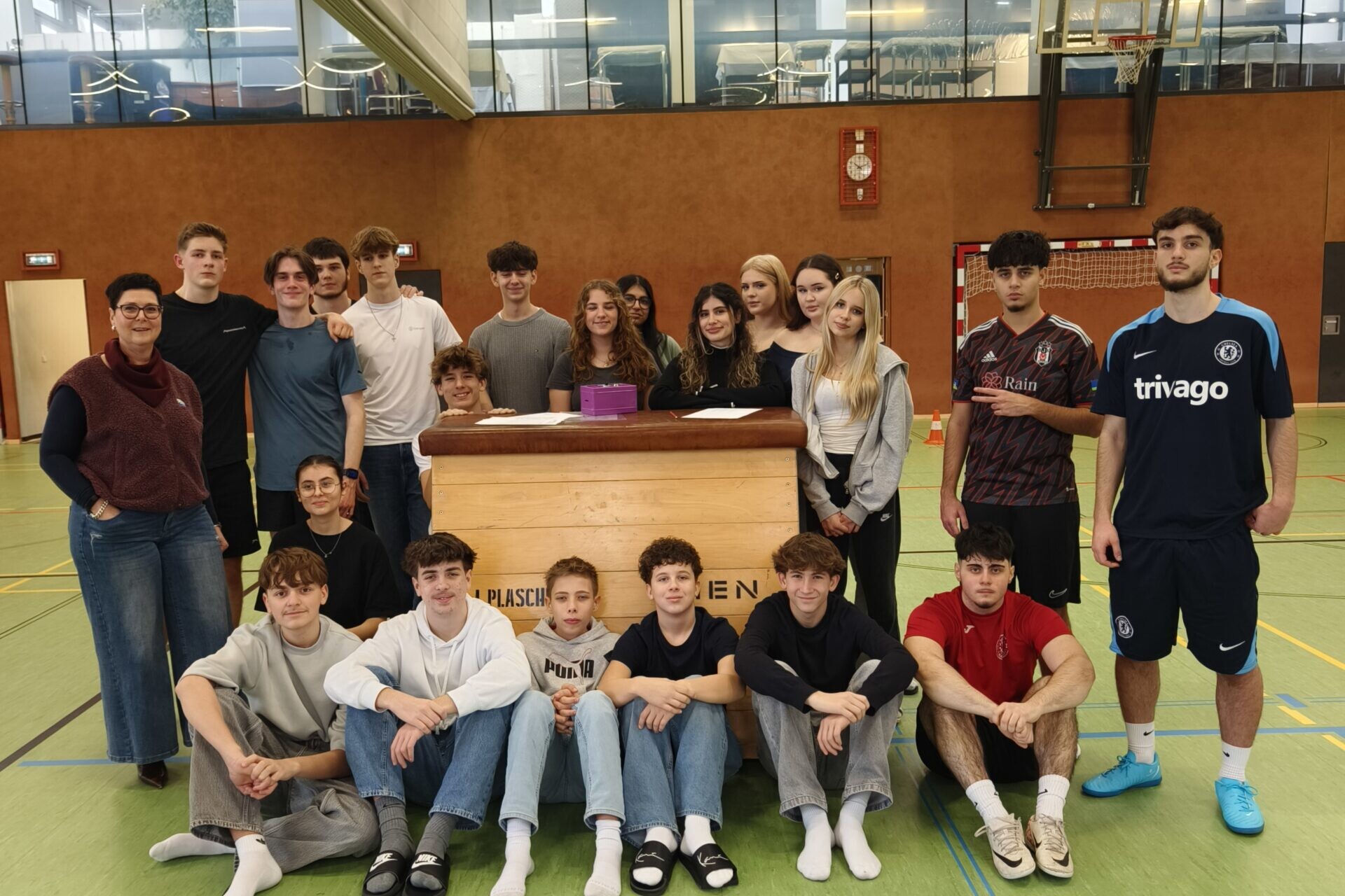 21 Schüler*innen gemeinsam mit Direktorin, direkt in die Kamera blickend, im Turnsaal.