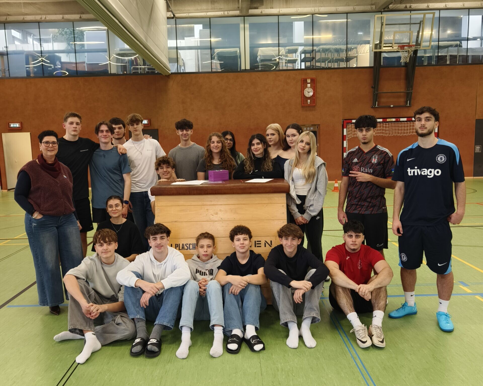 21 Schüler*innen gemeinsam mit Direktorin, direkt in die Kamera blickend, im Turnsaal.