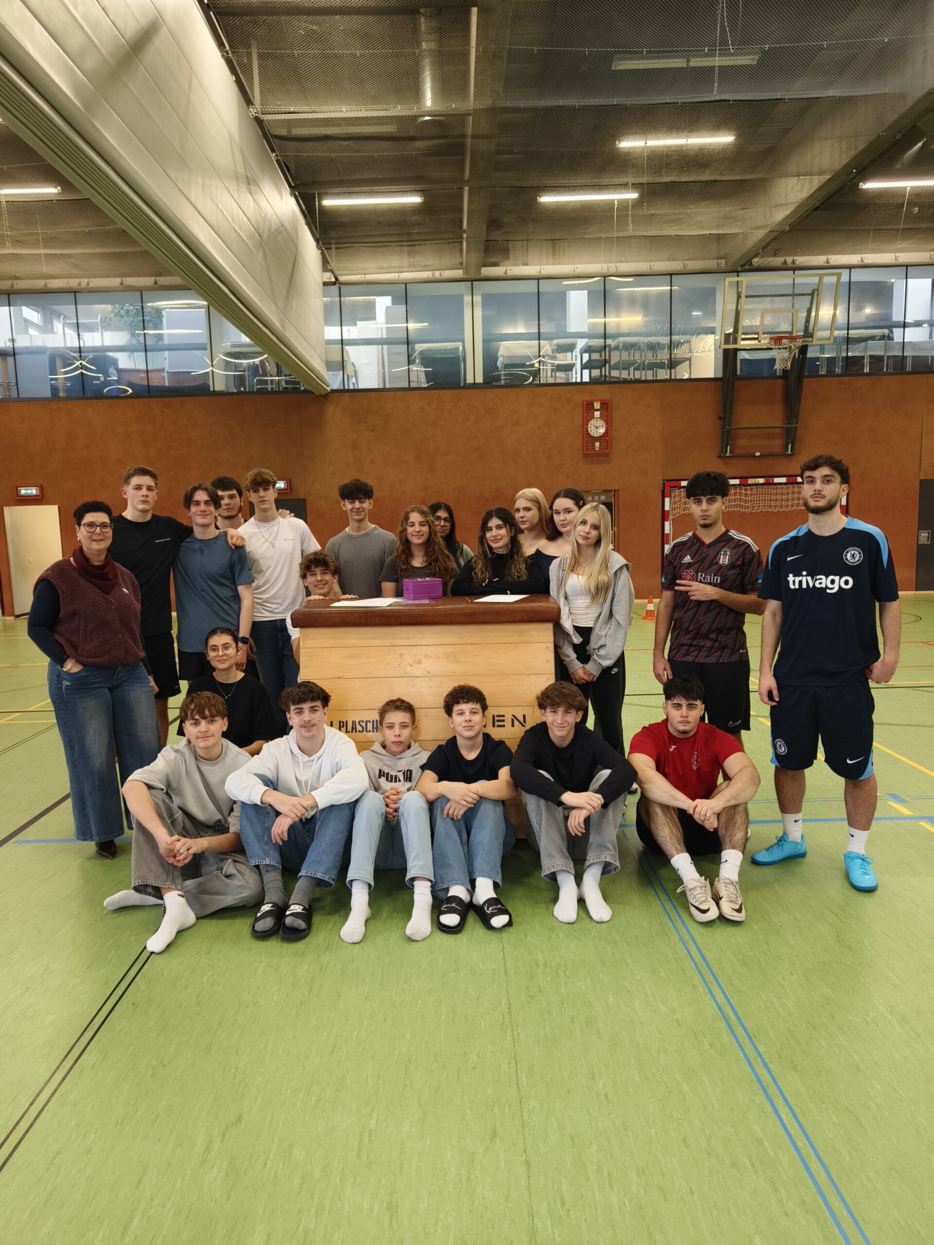 21 Schüler*innen gemeinsam mit Direktorin, direkt in die Kamera blickend, im Turnsaal.