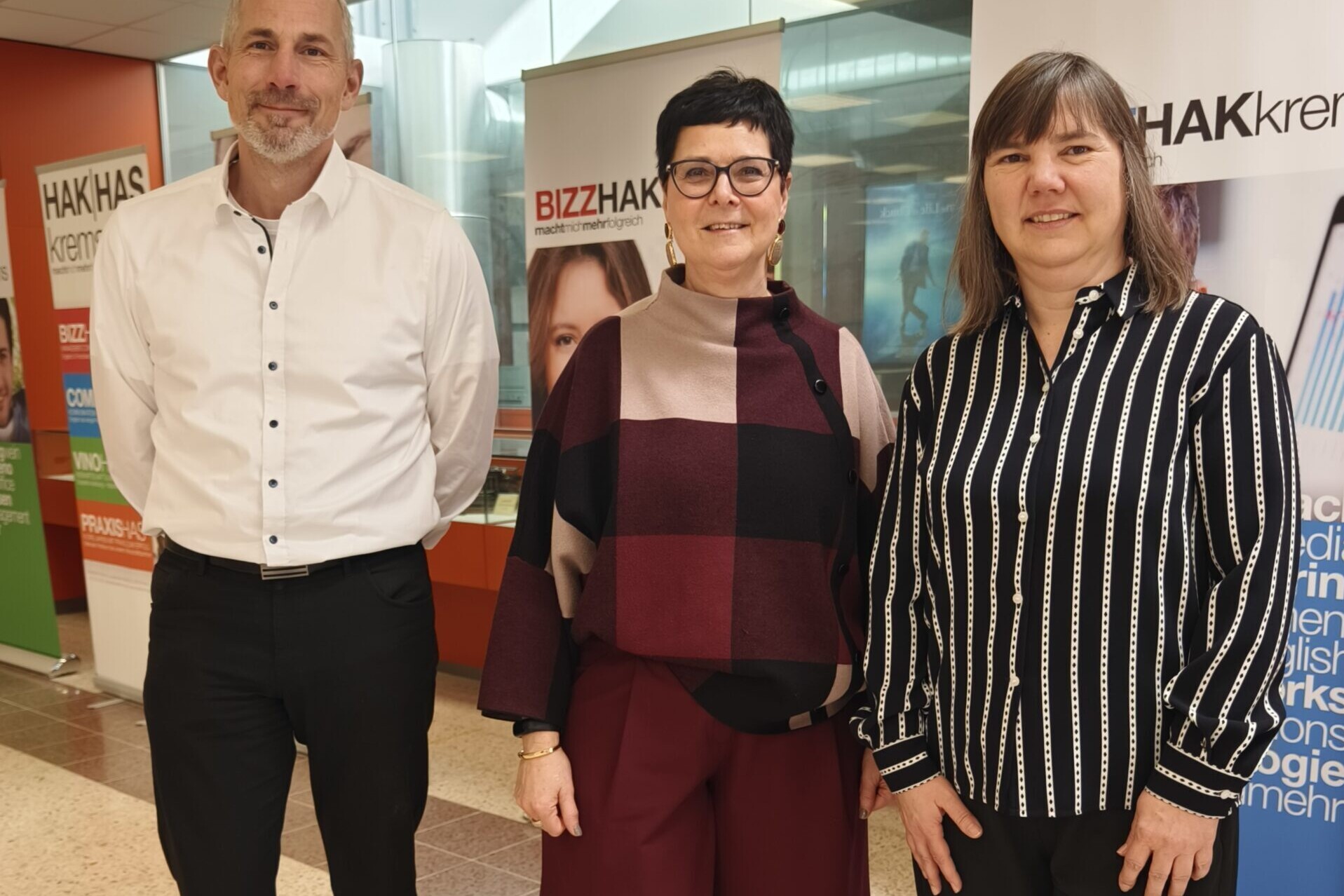 Zwei Frauen und ein Mann stehen lächelnd vor Banner der HAK Krems.
