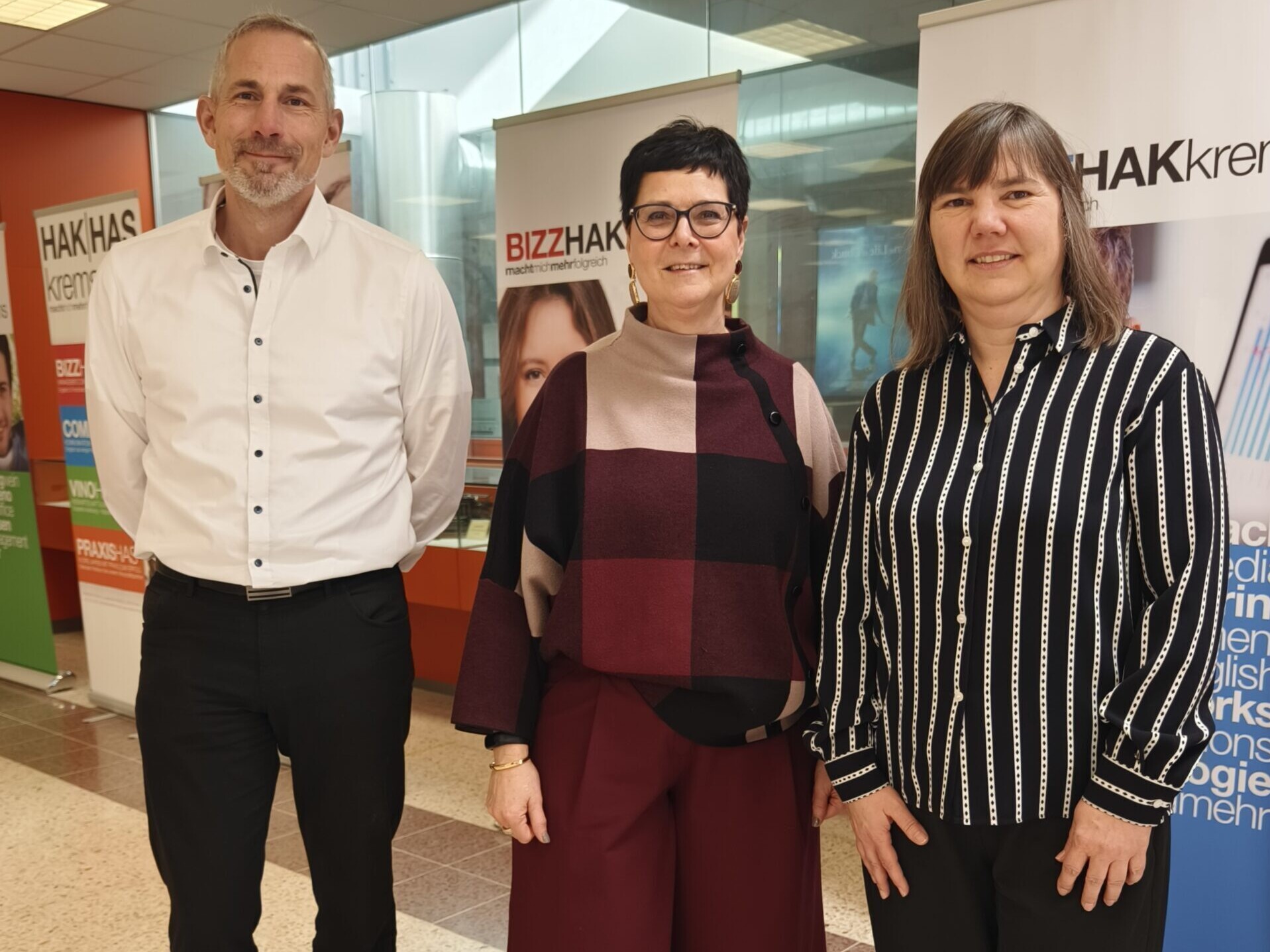 Zwei Frauen und ein Mann stehen lächelnd vor Banner der HAK Krems.