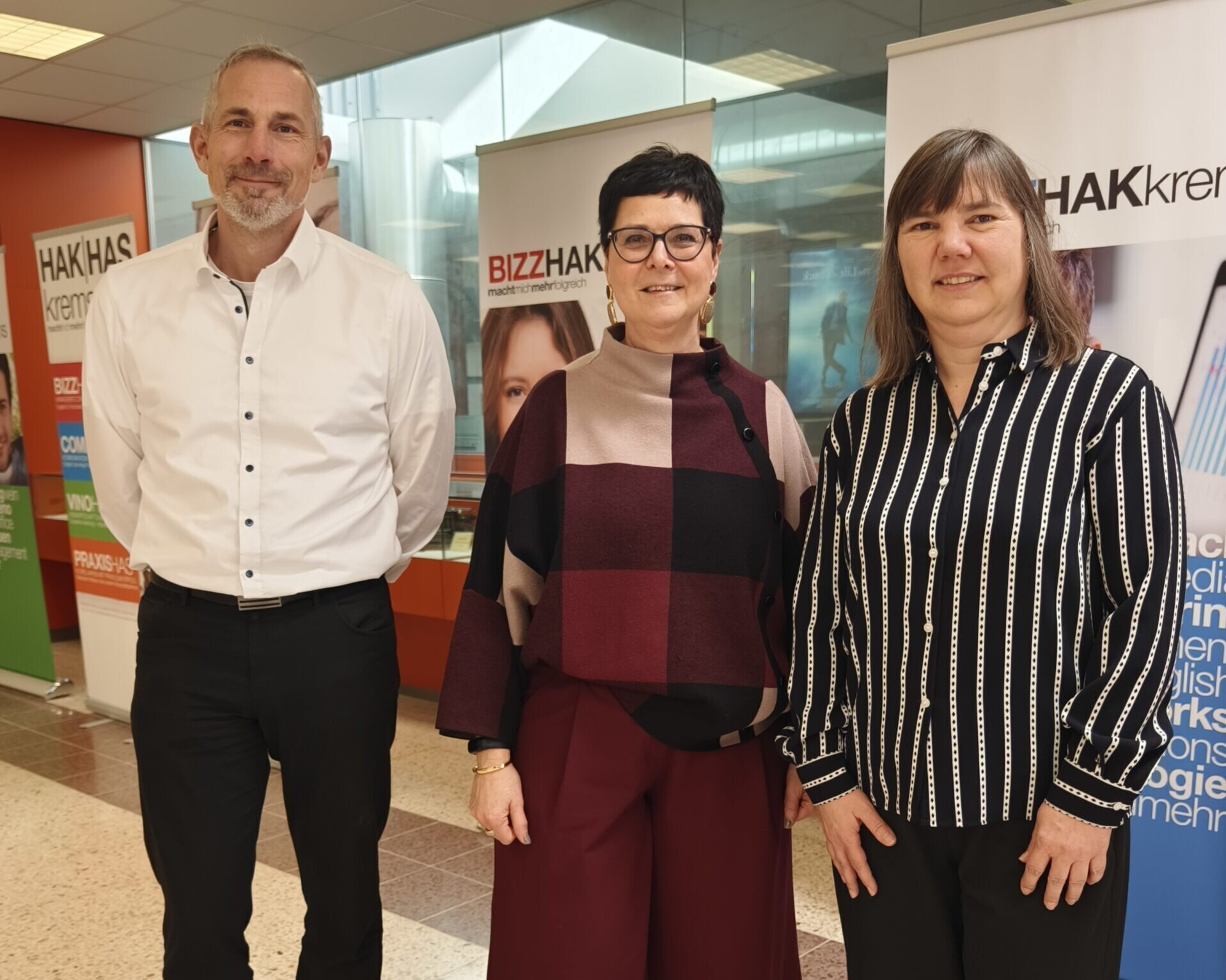 Zwei Frauen und ein Mann stehen lächelnd vor Banner der HAK Krems.