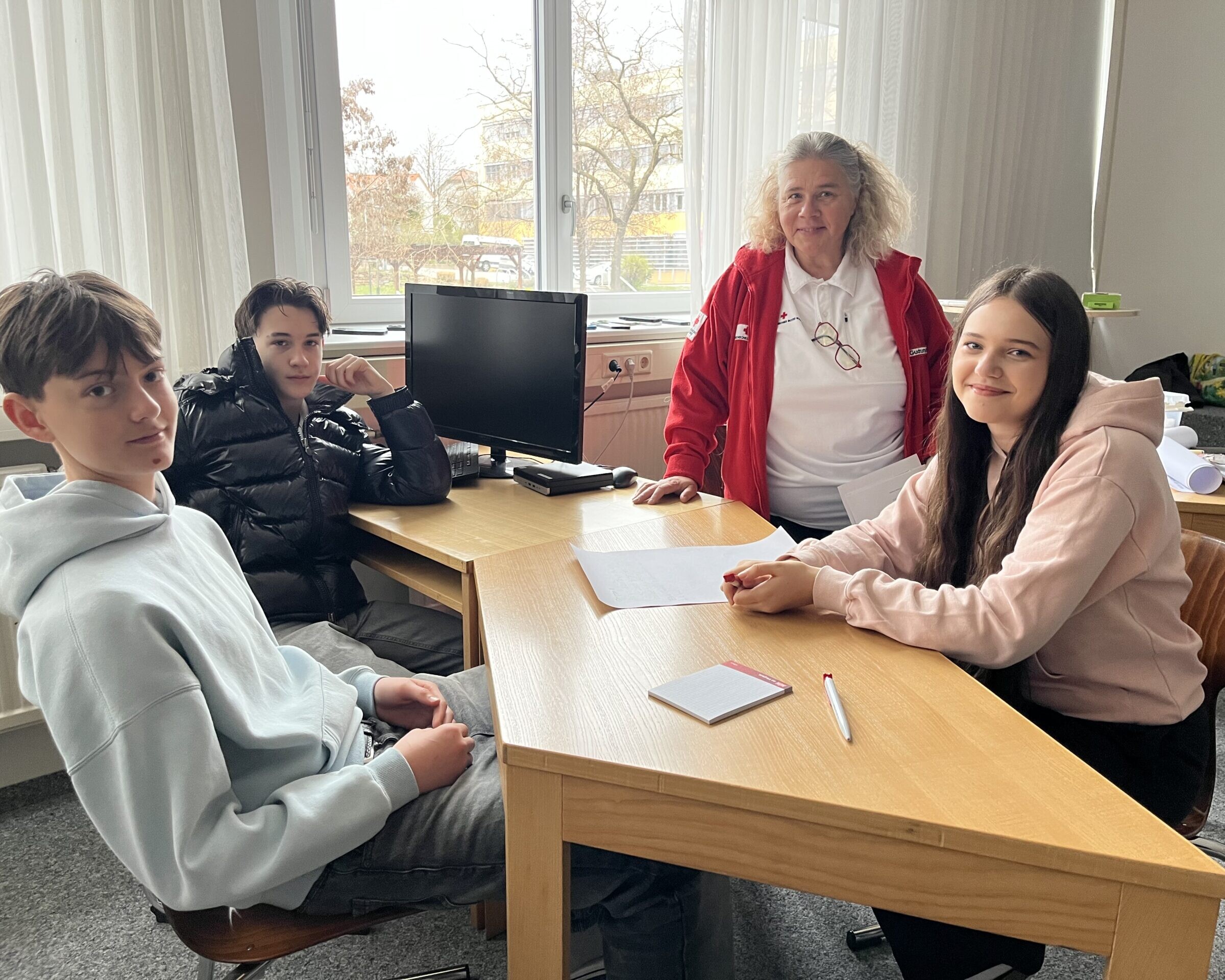 2 Schüler, 1 Schülerin und eine Frau vom Roten Kreuz sitzen rund um einen Tisch mit Zettel und einem Stift vor ihnen.