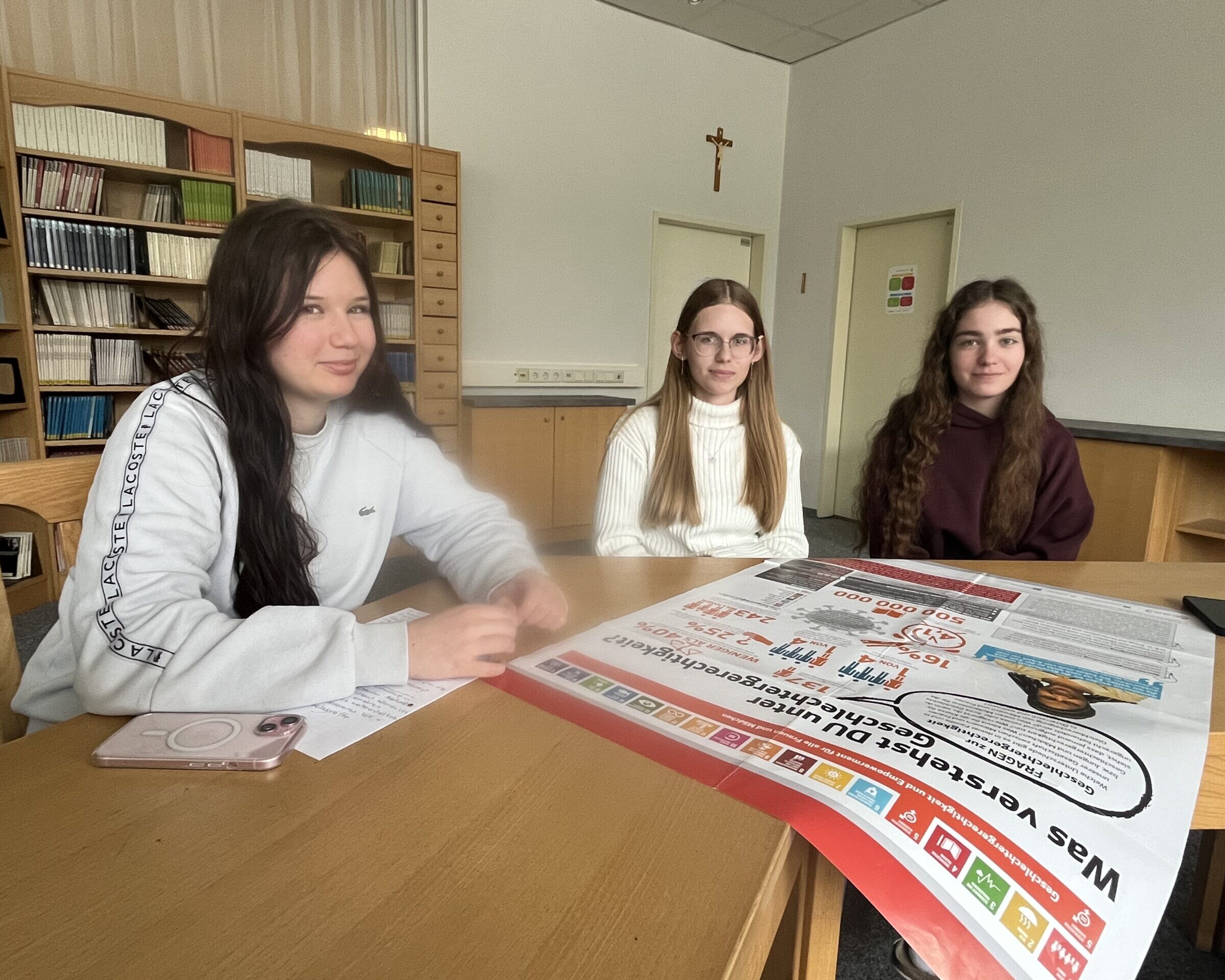 3 Schülerinnen sitzen bei einem Tisch, darauf liegt 1 NGO Plakat.