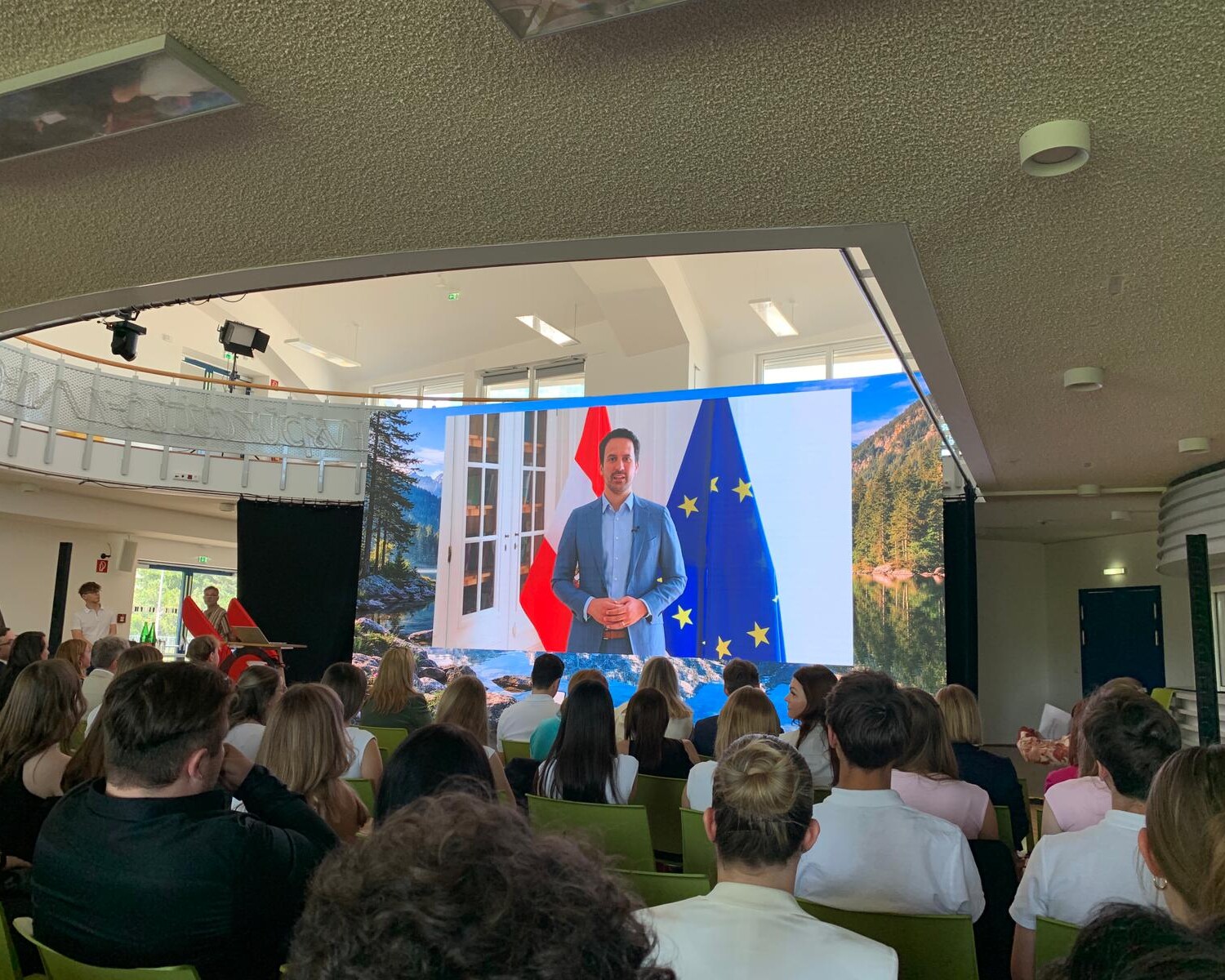 Publikum sitzt in einem Veranstaltungssaal und blickt auf eine große Leinwand, auf der eine Videobotschaft vor österreichischer und europäischer Flagge gezeigt wird.