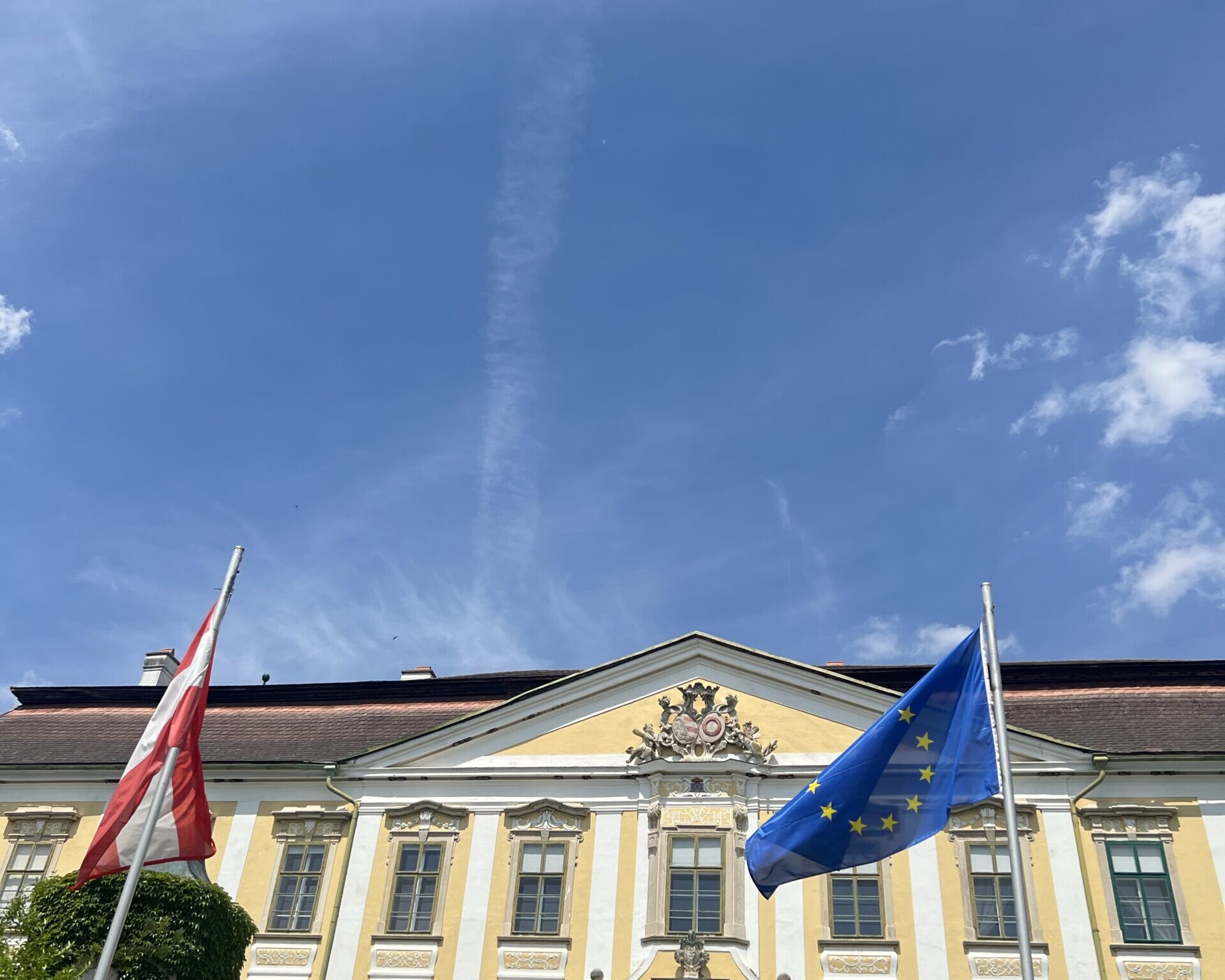 Ein historisches Gebäude mit gelb-weißer Fassade, davor die österreichische und die europäische Flagge unter blauem Himmel.