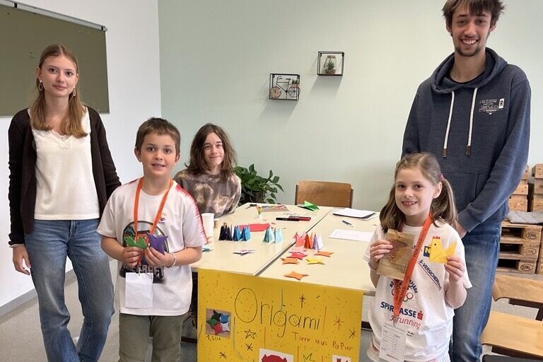 3 Schüler:innen und 2 Volkschulschüler:innen stehen rund um einen Tisch mit Origami darauf.