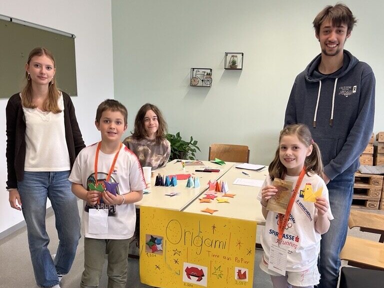 3 Schüler:innen und 2 Volkschulschüler:innen stehen rund um einen Tisch mit Origami darauf.