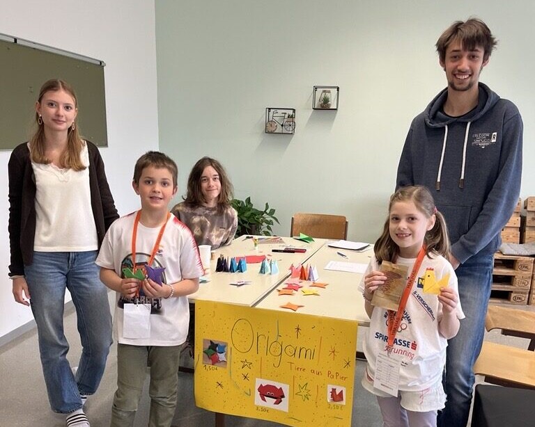 3 Schüler:innen und 2 Volkschulschüler:innen stehen rund um einen Tisch mit Origami darauf.