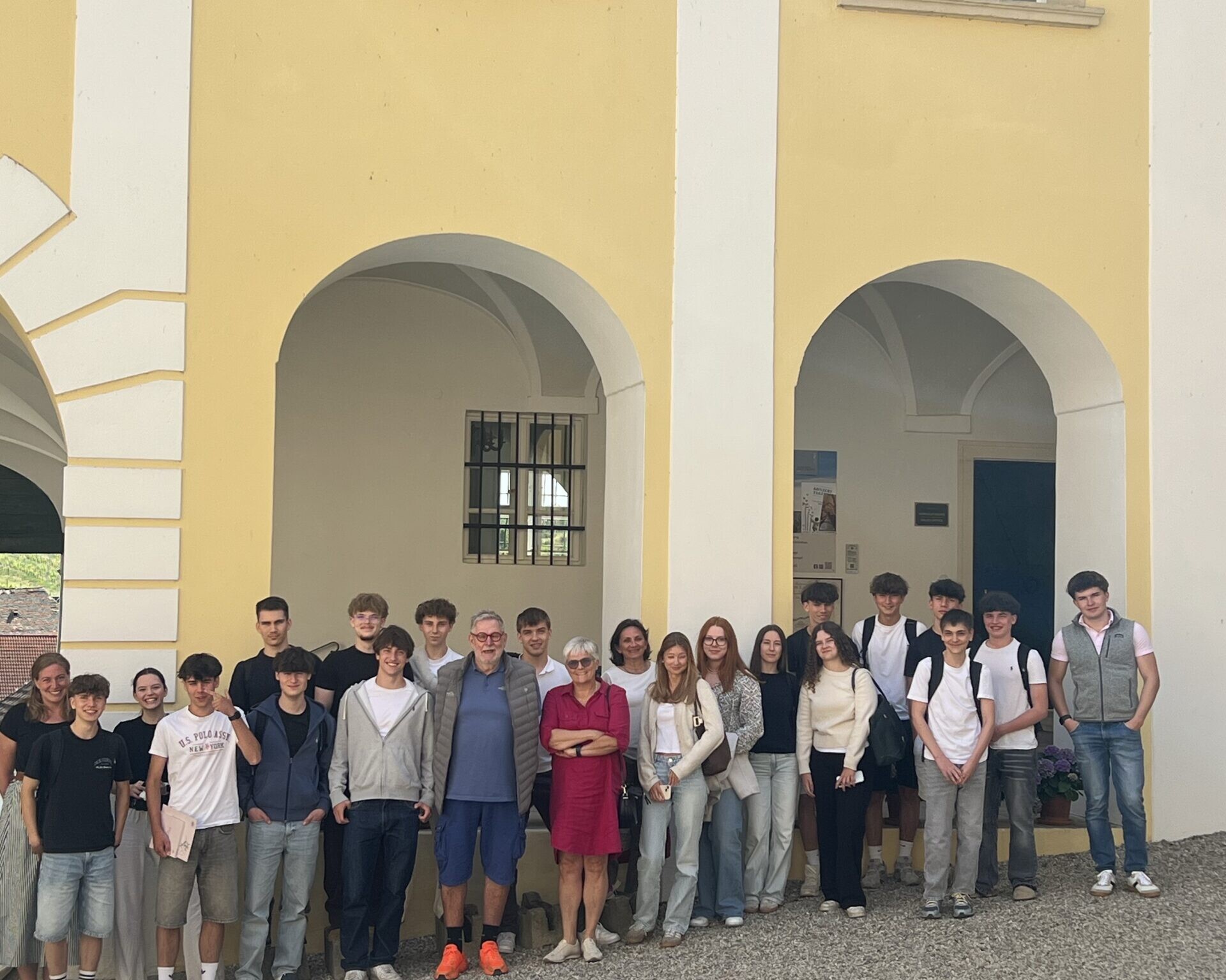 Eine Gruppe von Schülerinnen und Schülern posiert gemeinsam vor einem historischen Gebäude mit hohen Bögen und gelber Fassade.