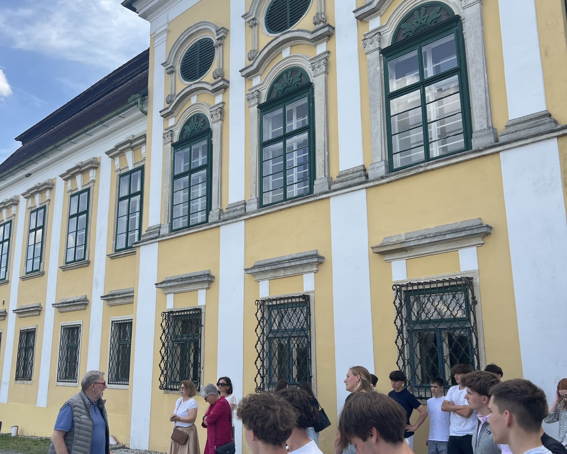 Eine Gruppe von Menschen steht vor einem gelb-weißen historischen Gebäude mit hohen Fenstern, während eine Person offenbar eine Führung oder Erklärung gibt.