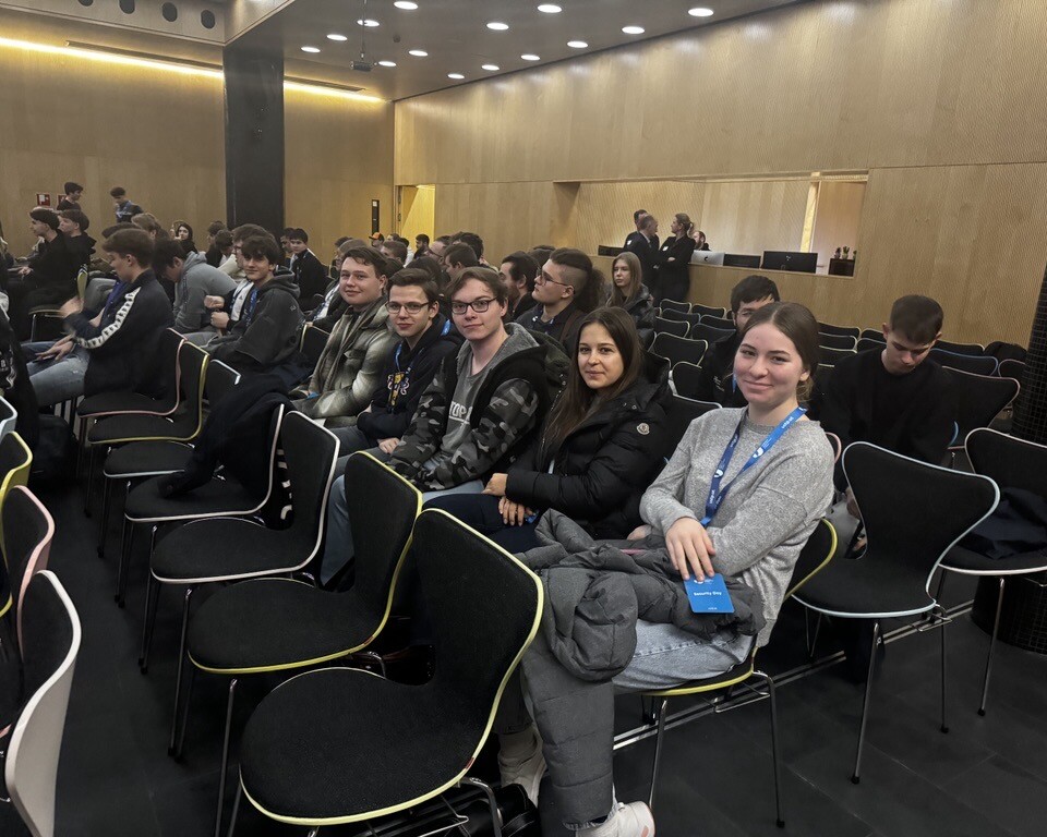 Eine Gruppe junger Personen sitzt in mehreren Reihen von Stühlen in einem modernen Veranstaltungssaal und schaut lächelnd in Richtung Kamera.