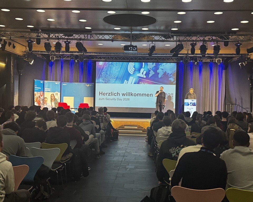Viele Menschen sitzen in einem Veranstaltungssaal und hören zwei Personen auf einer Bühne zu, während auf einer großen Leinwand eine Begrüßungsfolie angezeigt wird.