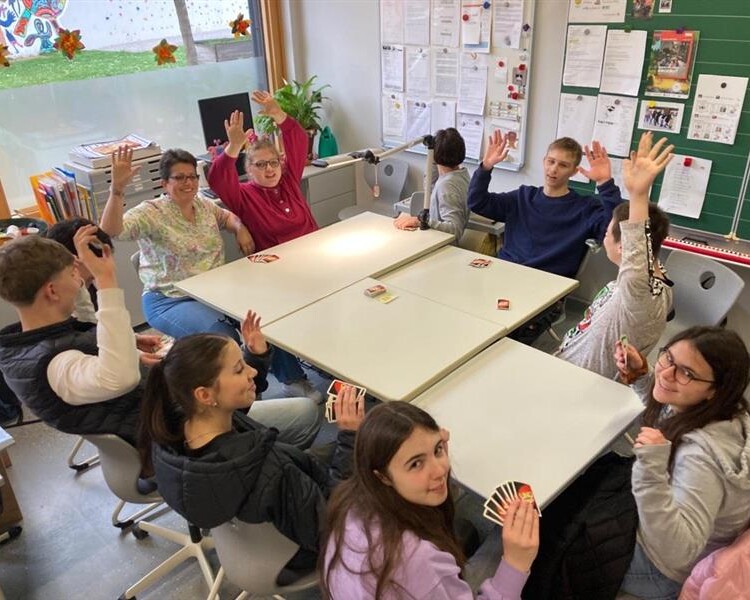 Mehrere Schüler und eine Erwachsene Person sitzen um zusammengestellte Tische, heben die Hände und halten Spielkarten, während sie in einem hellen Klassenraum spielen.